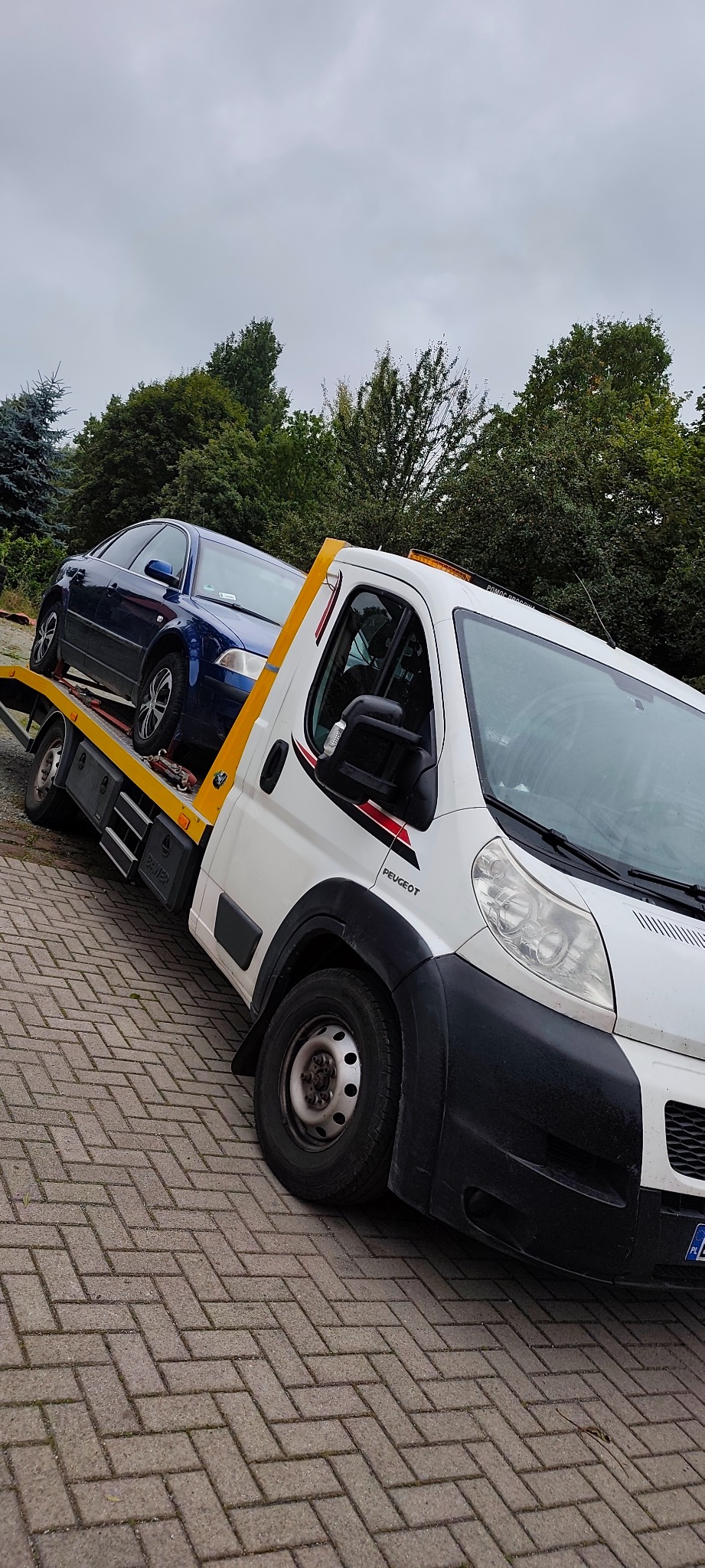 Laweta Peugeot z niebieskim autem osobowym na platformie, zaparkowana na brukowanym placu, z drzewami w tle i pochmurnym niebem. Pomoc drogowa.