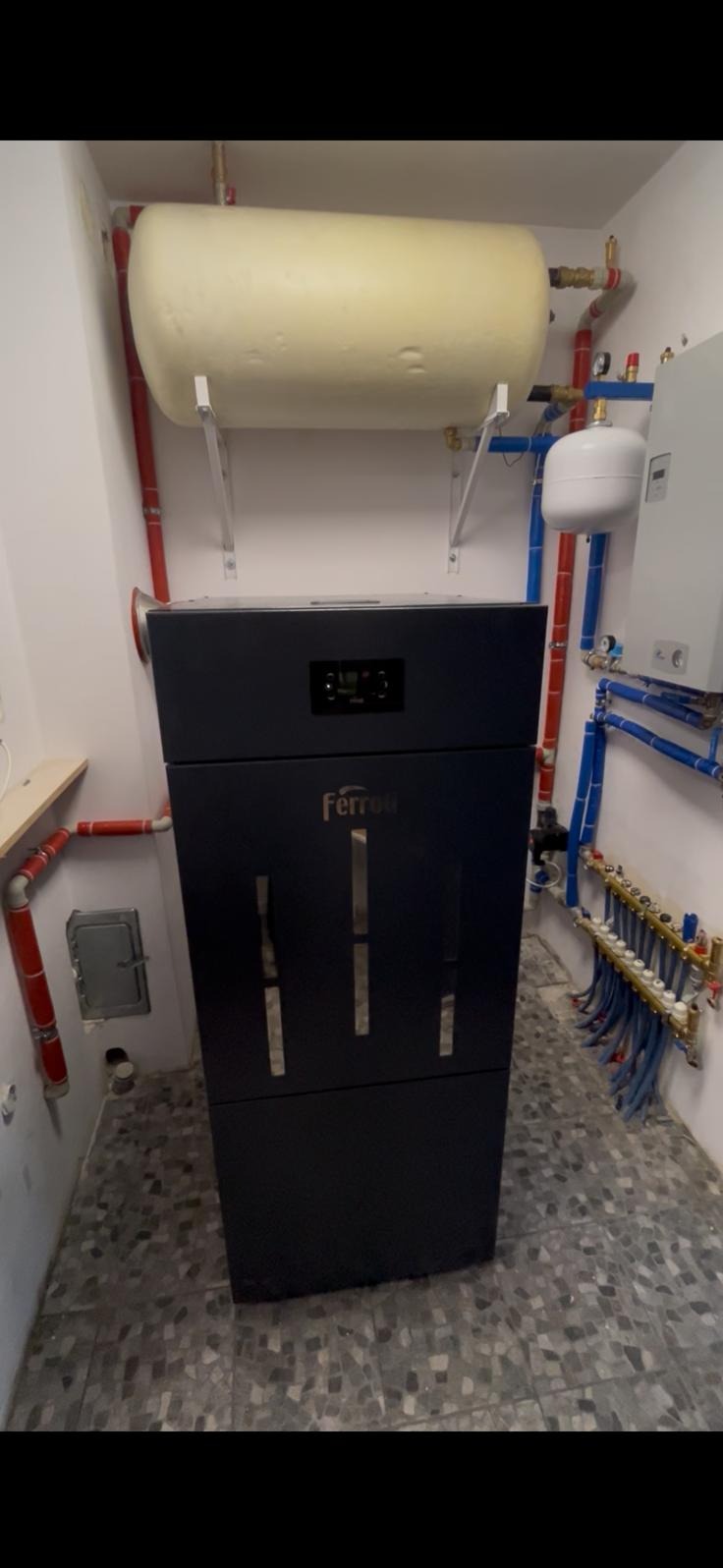 Nowoczesny piec CO marki Ferroli w kotłowni z instalacją grzewczą i zbiornikiem buforowym. Widoczne rury w kolorach czerwonym i niebieskim oraz szara, mozaikowa podłoga.