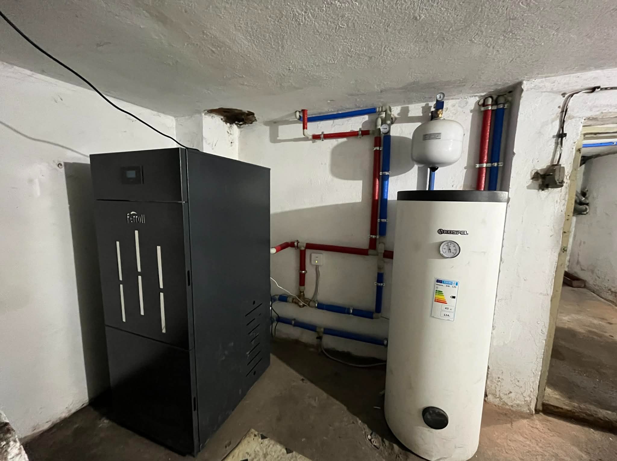 Nowoczesny piec Ferroli obok białego zasobnika Kospel SW-120, z widoczną instalacją hydrauliczną w kolorach czerwonym i niebieskim, w surowym wnętrzu kotłowni.