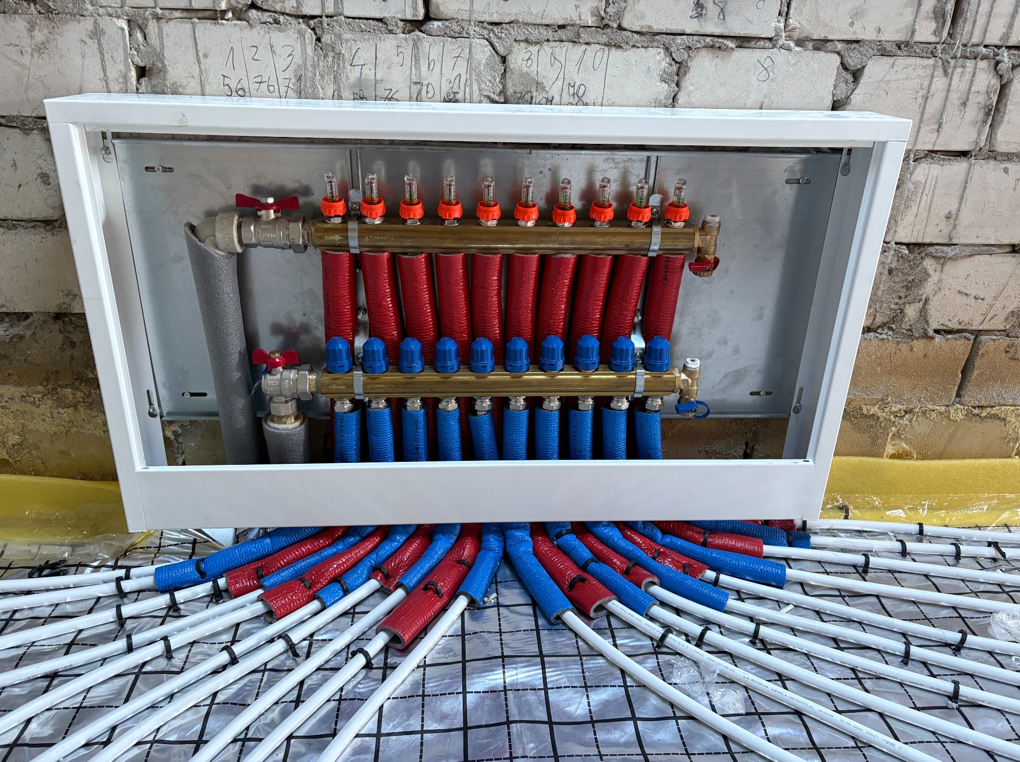 Rozdzielacz ogrzewania podłogowego w białej szafce, z rurami w izolacji czerwonej i niebieskiej, ułożonymi na folii z rastrem, na tle ściany z cegły.