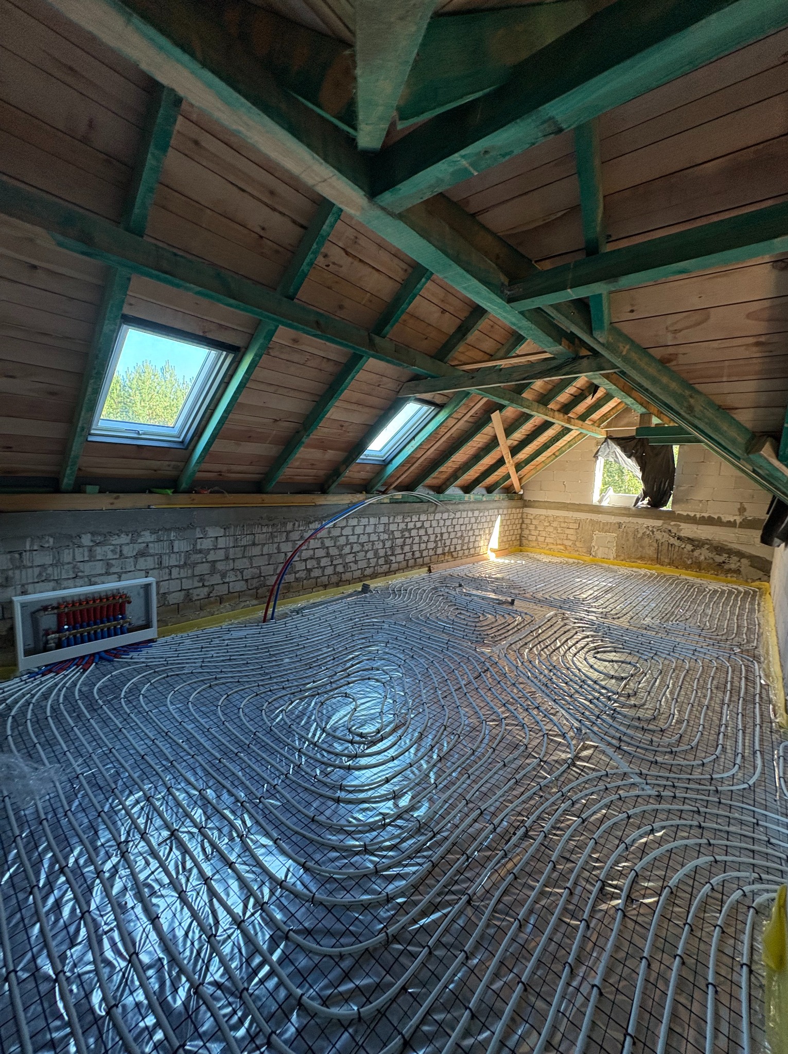Ogrzewanie podłogowe w trakcie montażu na poddaszu: rury ułożone na folii, widoczny rozdzielacz, okna dachowe i drewniana konstrukcja dachu.