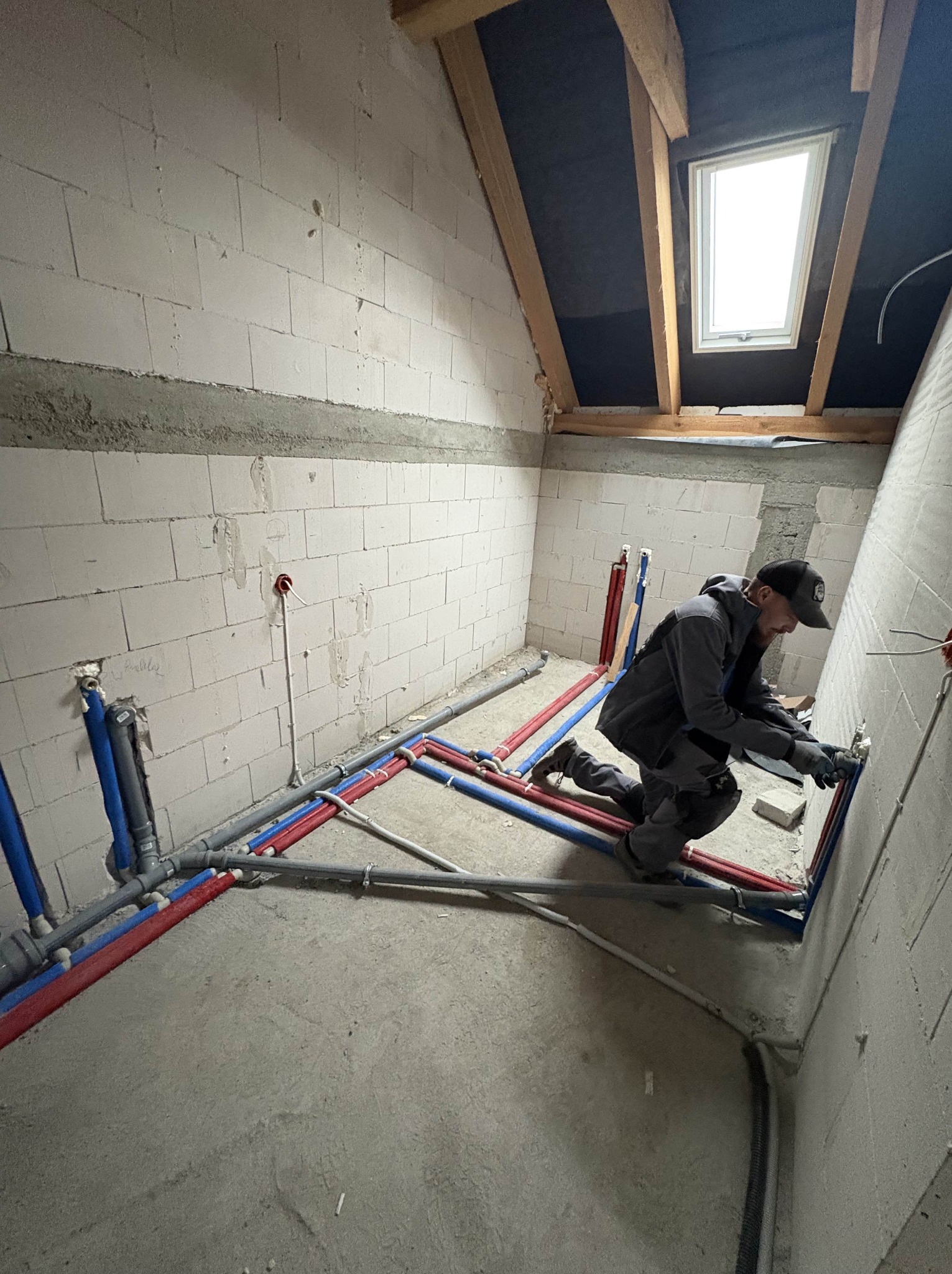 Hydraulik w trakcie montażu instalacji wodnej w surowym wnętrzu poddasza. Widoczne rury w kolorach niebieskim i czerwonym oraz okno dachowe.
