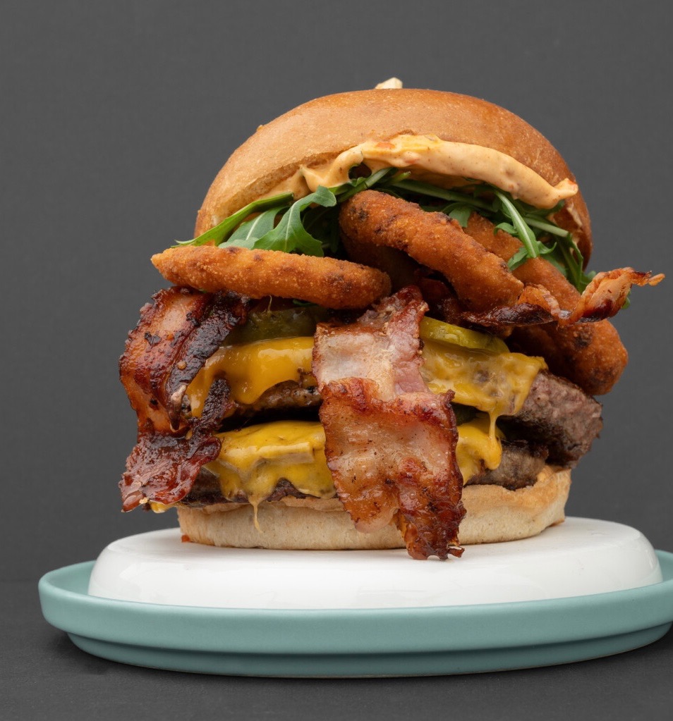 Apetyczny, dwuwarstwowy burger z bekonem, serem, krążkami cebulowymi i rukolą, podany na talerzu z niebieskim rantem na ciemnoszarym tle. Idealny na catering.