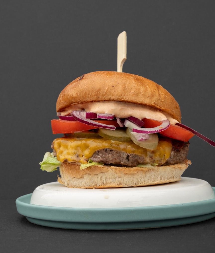 Apetyczny burger z wołowiną, serem cheddar, pomidorem, czerwoną cebulą i sosem, umieszczony na talerzu z turkusowym brzegiem, na ciemnym tle.
