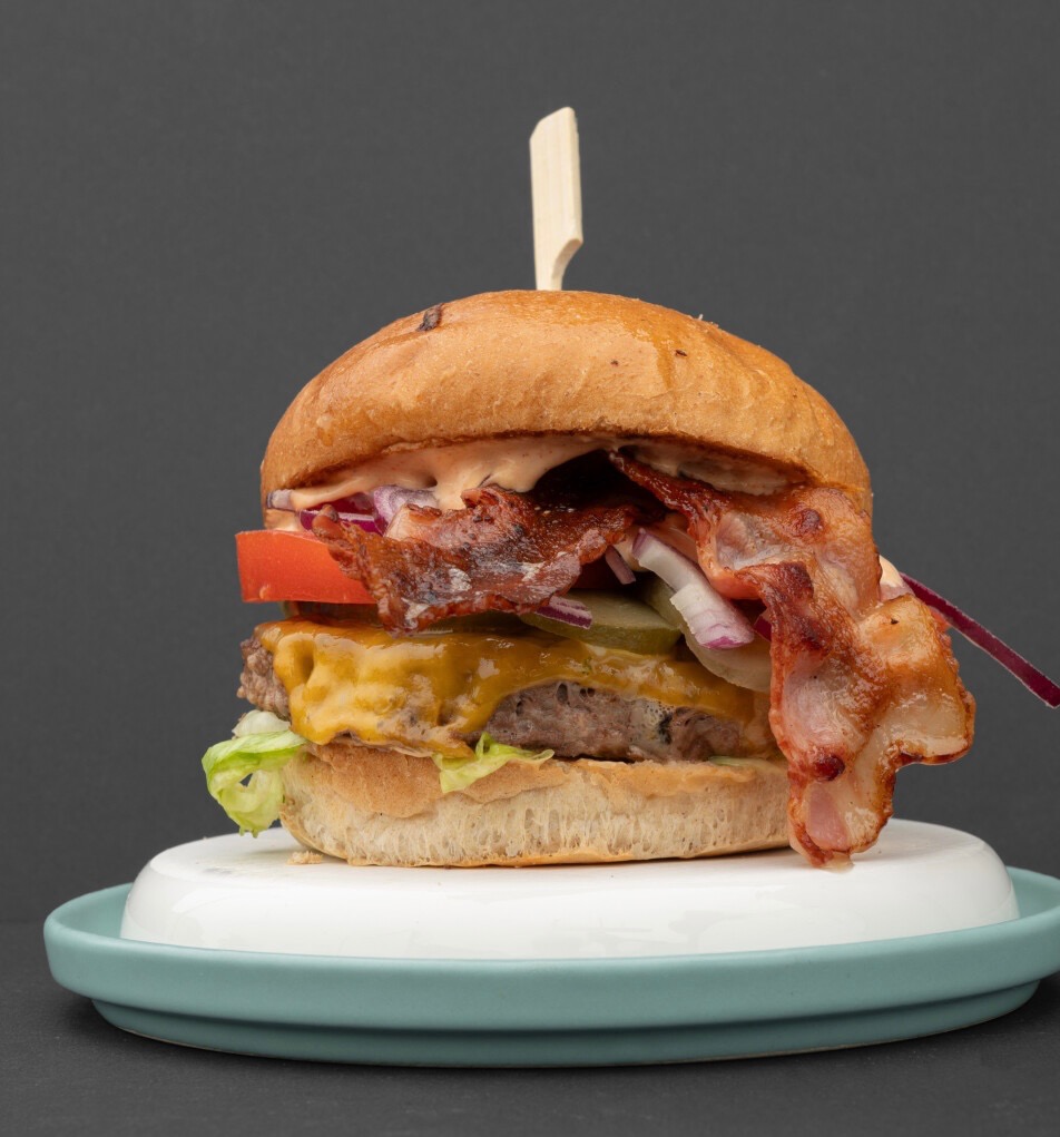 Apetyczny burger z bekonem, serem i warzywami na białym talerzu z niebieskim rantem, na ciemnoszarym tle. Widoczny w całości, z bliska.