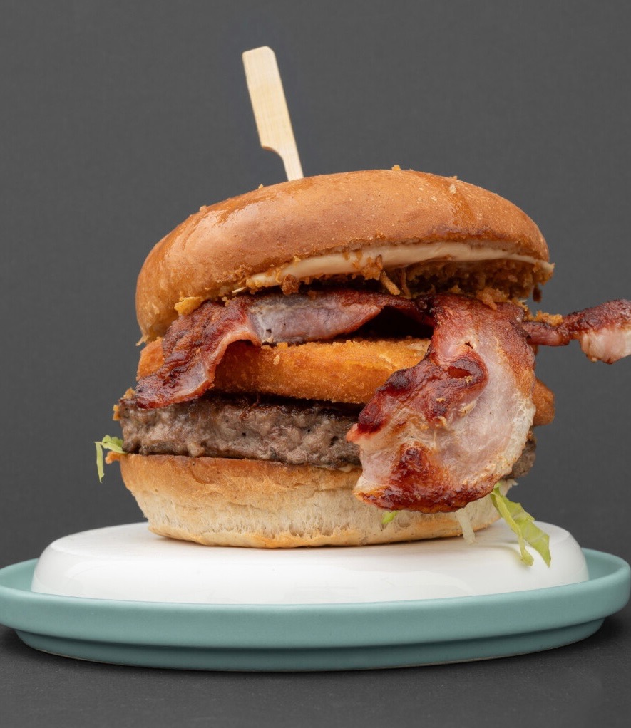 Apetyczny, wysoki burger z bekonem i krążkiem cebulowym, na białym talerzu z turkusowym brzegiem, na szarym tle. Widoczny drewniany patyczek.