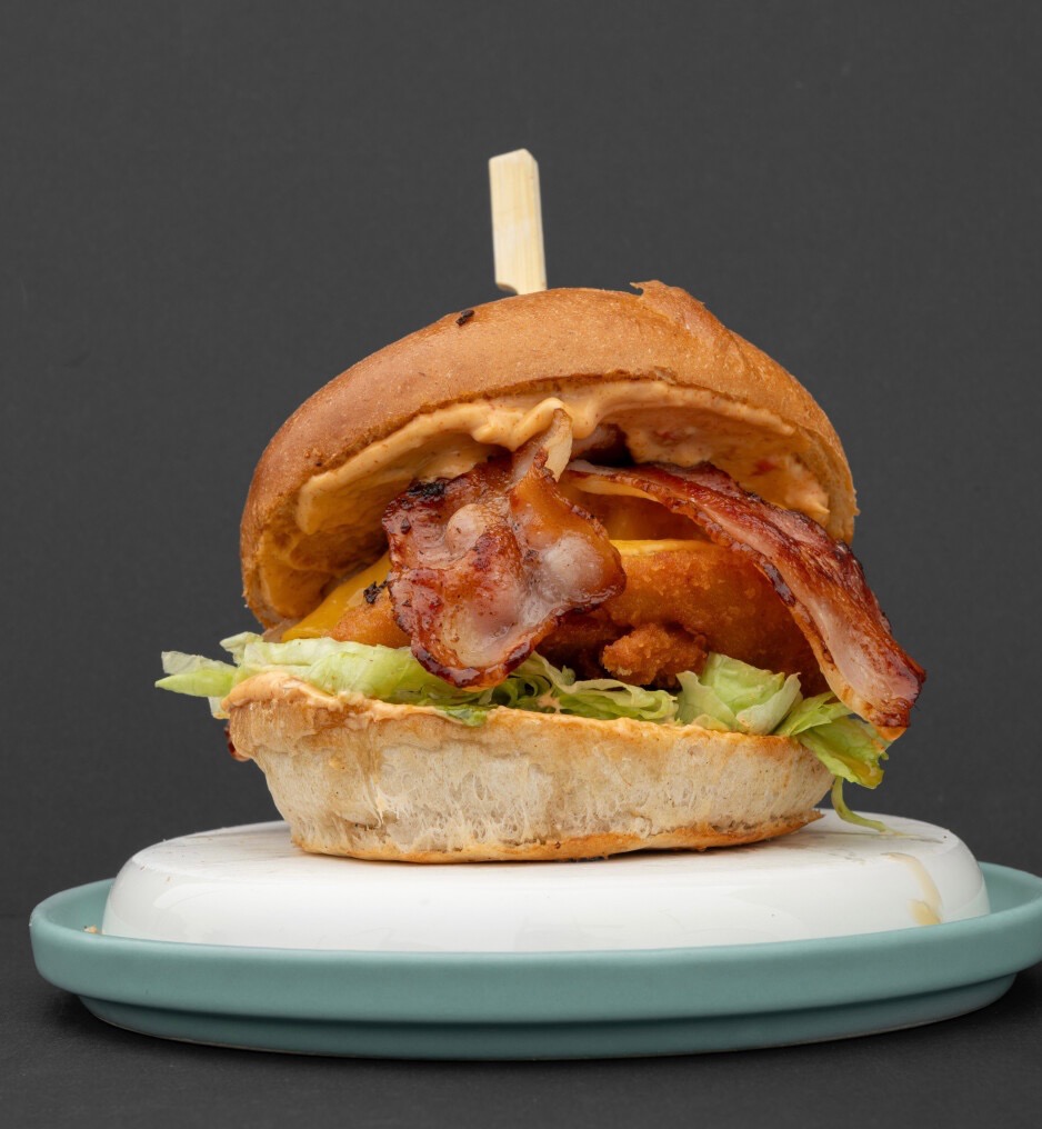 Apetyczny burger z bekonem, serem i sosem na talerzu z turkusowym brzegiem, na tle ciemnoszarego tła. Widoczne warstwy sałaty i panierowanego mięsa.