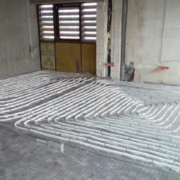 Białe rury ogrzewania podłogowego ułożone na siatce w surowym wnętrzu, widoczne okno z żaluzjami i fragmenty ścian w stanie deweloperskim.