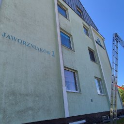 SLAWOMIREX SŁAWOMIR SKOWROŃSKI - Elewacja budynku z widocznymi zabrudzeniami przed czyszczeniem, drabina oparta o ścianę. Osoba w żółtej kurtce przygotowuje się do pracy. Błękitne niebo w tle.