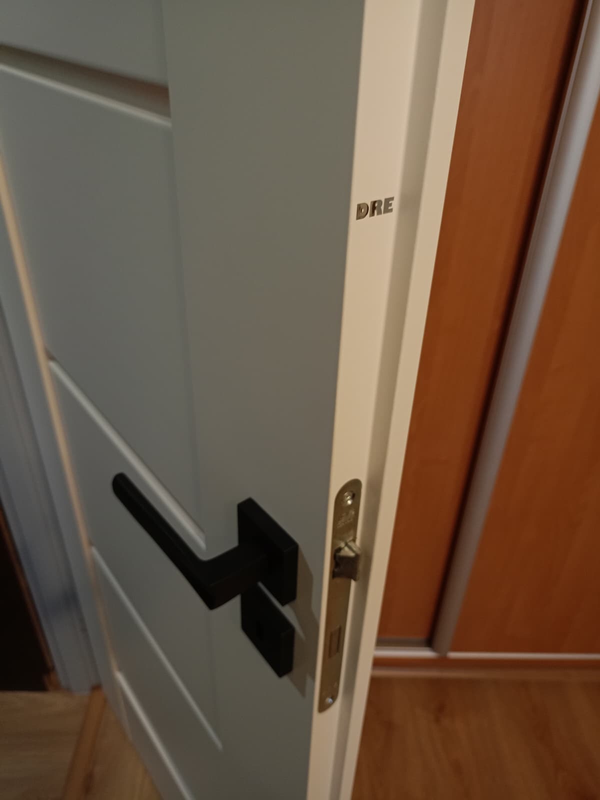 Białe drzwi wewnętrzne z czarną klamką i widocznym zamkiem, marki DRE, na tle fragmentu szafy. Ujęcie z bliska, z boku, ukazujące detale wykończenia.