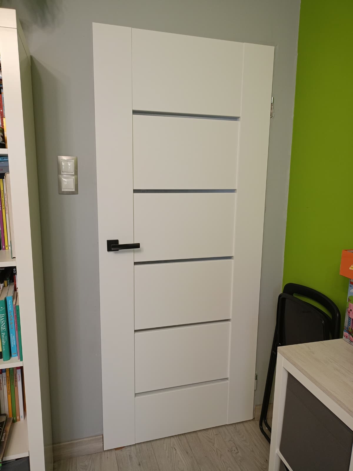 Białe drzwi wewnętrzne z poziomymi frezami i czarną klamką, widoczne na tle szarej ściany z włącznikiem i fragmentem regału z książkami.