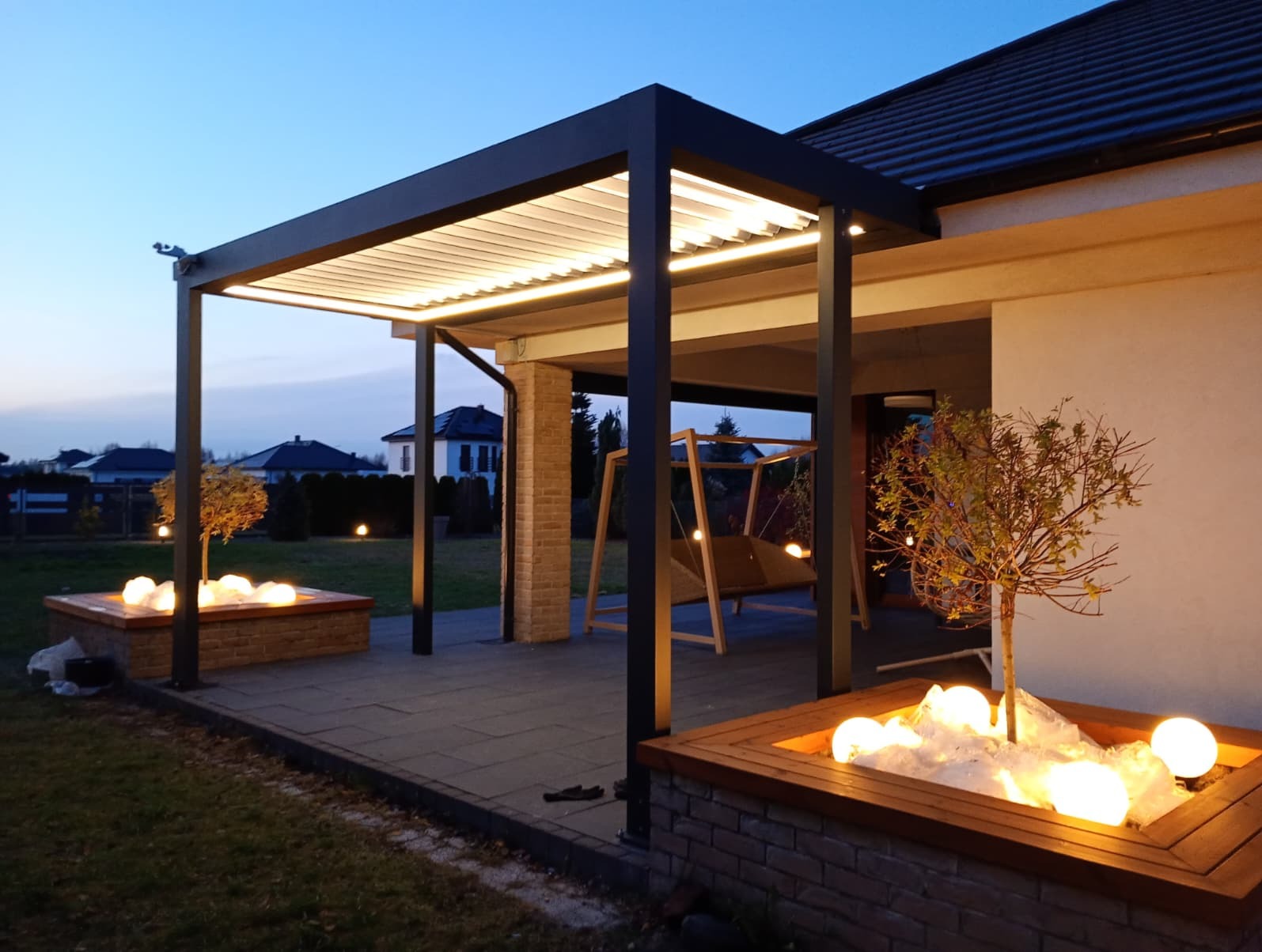 Nowoczesna, czarna pergola metalowa z oświetleniem LED na tarasie. Widoczne donice z podświetlonymi kulami i huśtawka ogrodowa w tle, wieczorny krajobraz.