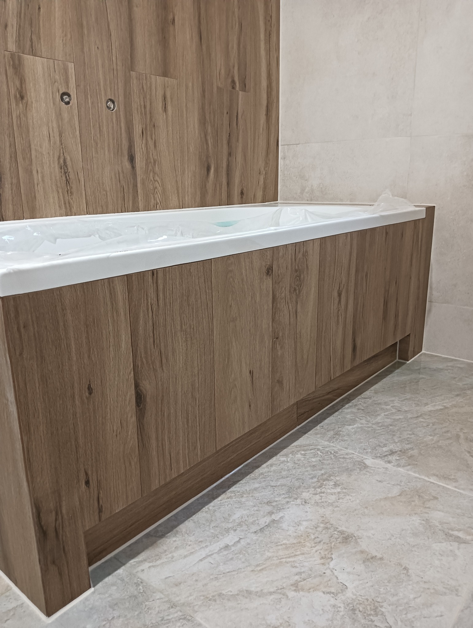 Obudowana wanna z drewnopodobną okładziną i jasnymi płytkami. Nowoczesna łazienka z subtelnym oświetleniem i podłogą z płytek imitujących kamień.