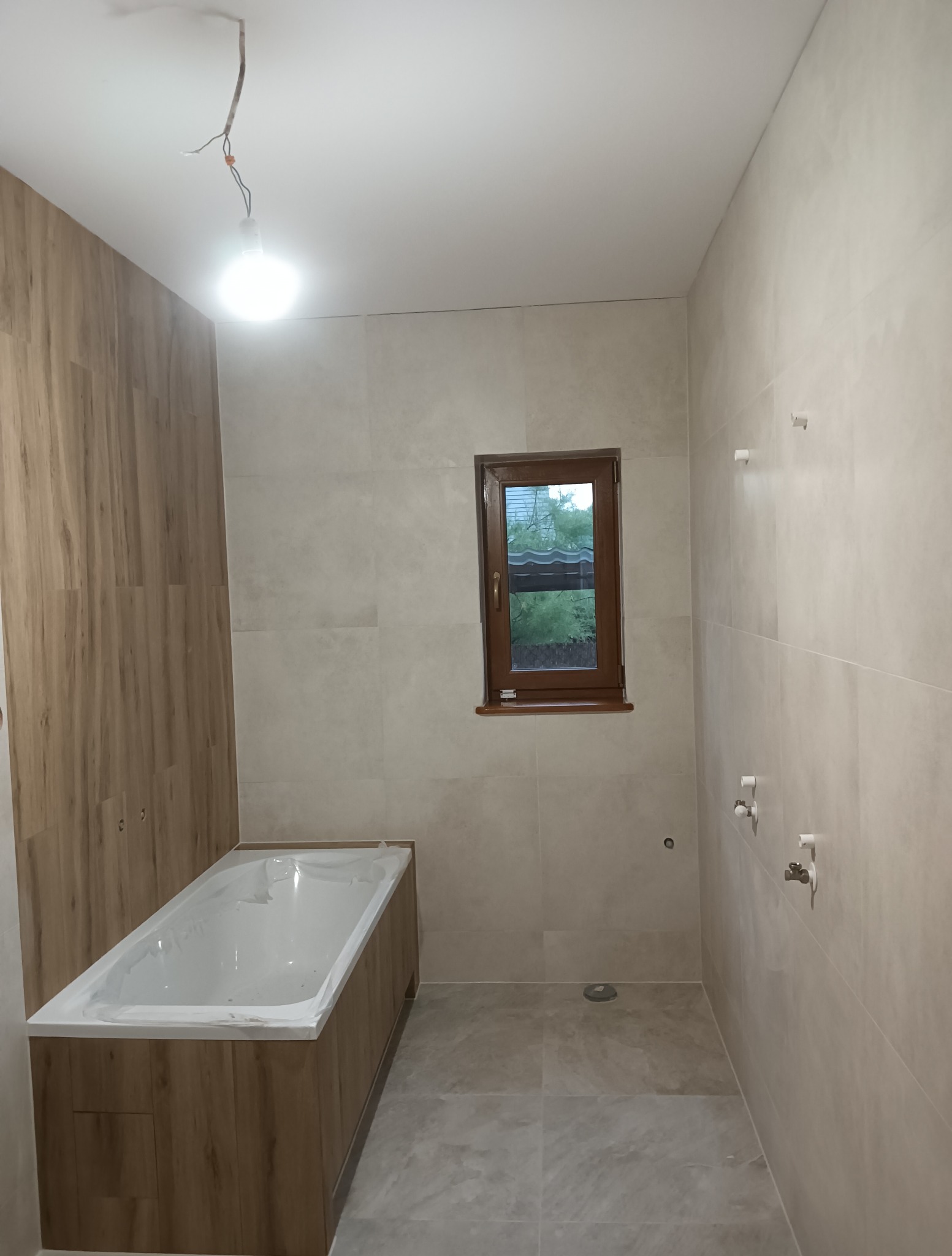 Wykończona łazienka z wanną w obudowie z drewnopodobnych paneli i beżowymi płytkami. Widoczne okno z brązową ramą i wystające przewody oświetleniowe.