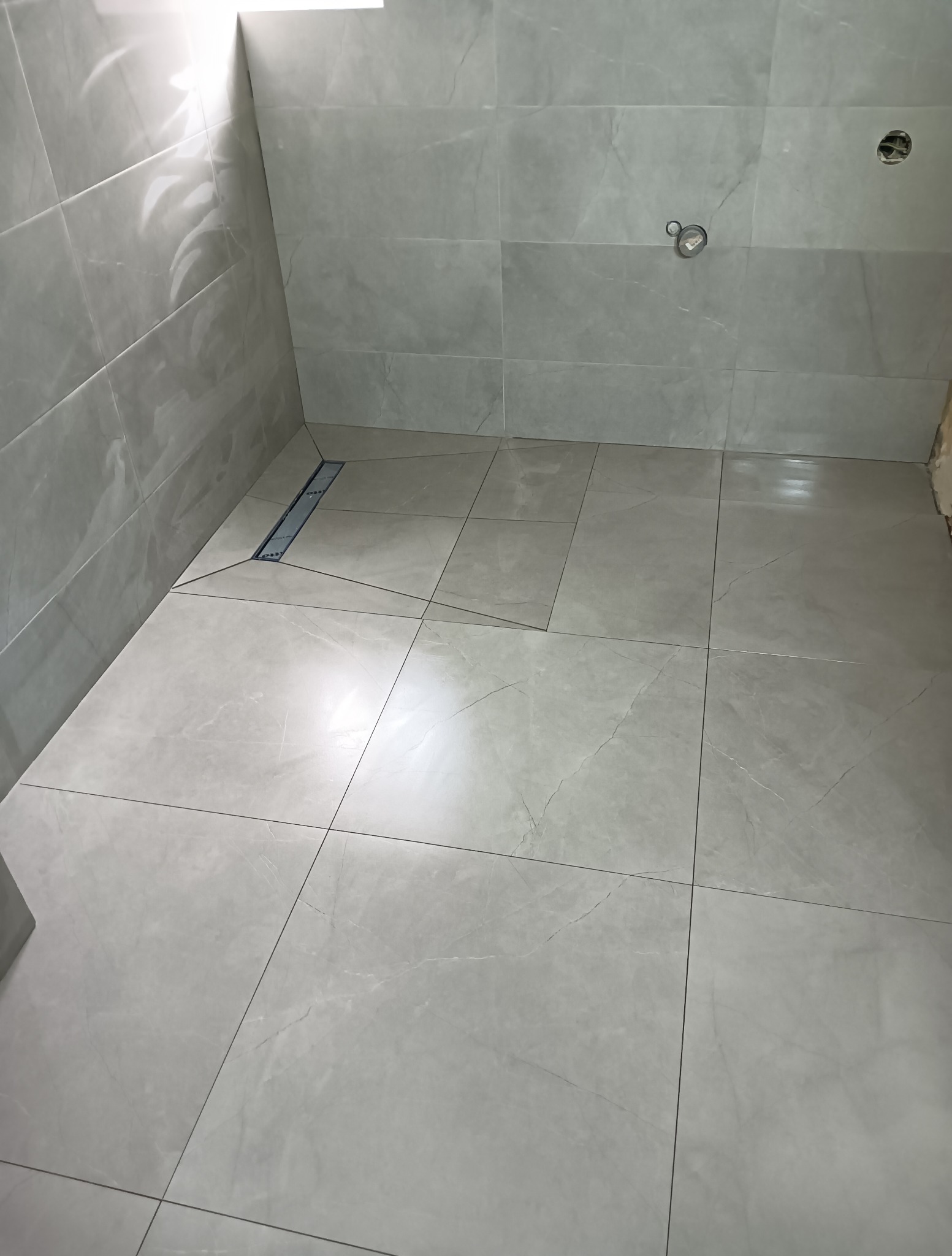 Jasnoszare, matowe płytki w łazience z odpływem liniowym. Widoczne otwory na armaturę. Minimalistyczny design, surowy klimat.