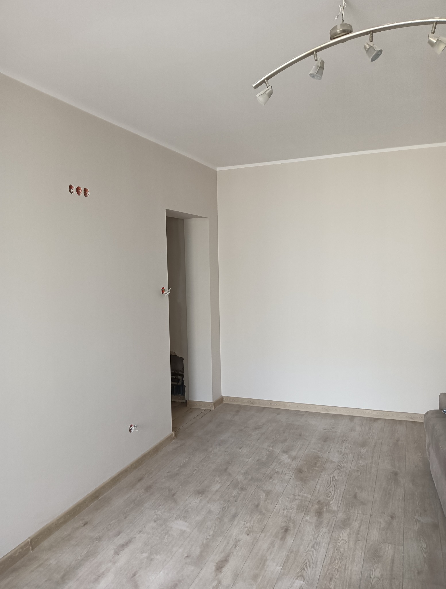 Jasne panele podłogowe w wyremontowanym pokoju z beżowymi ścianami. Widoczne puszki elektryczne i fragment sofy. Nowoczesne oświetlenie sufitowe.