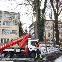 P.U.H. ALPE - EDWARD IDZI-JAROMIJ - Pracownik w odblaskowej odzieży przy podnośniku koszowym na tle miejskiego krajobrazu i maszyny do rozdrabniania gałęzi. Wycinka drzew w przestrzeni miejskiej.