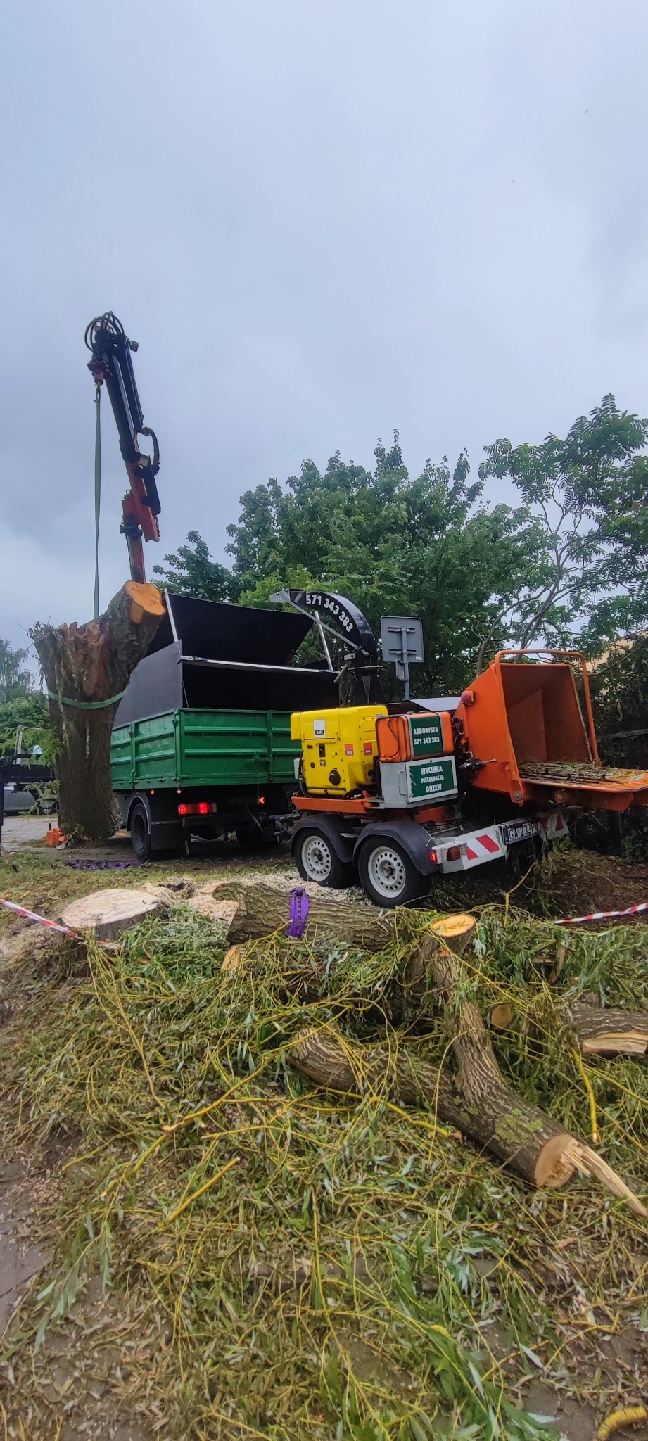 Wycinka drzewa z użyciem dźwigu i rębaka. Fragment pnia jest podnoszony do skrzyni ciężarówki, a obok rębakiem rozdrabniane są gałęzie. Praca w toku.