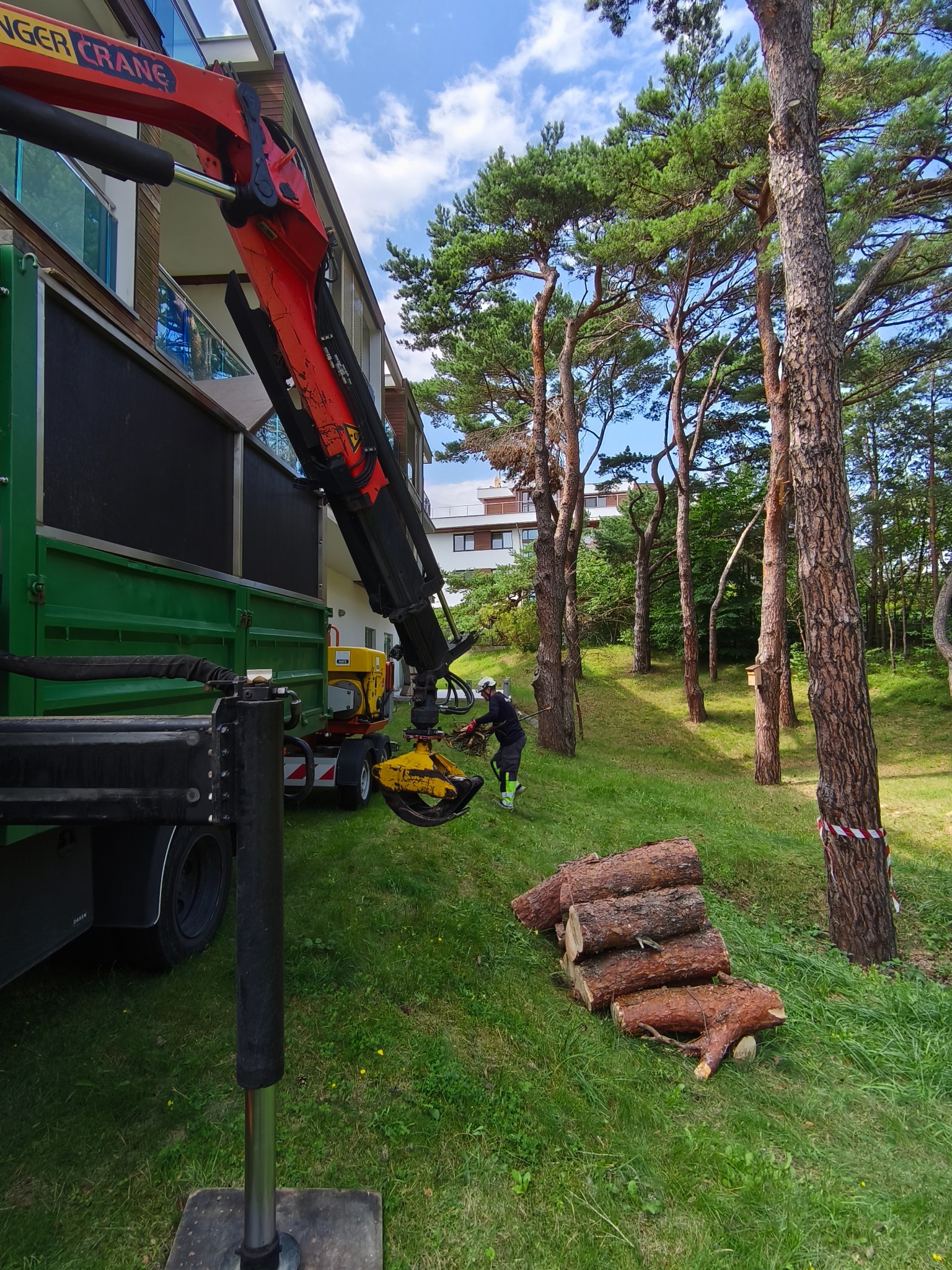 Praca przy wycince drzew: operator w kasku obsługuje dźwig z chwytakiem, obok leżą pocięte pnie, a w tle widoczny budynek i drzewa.