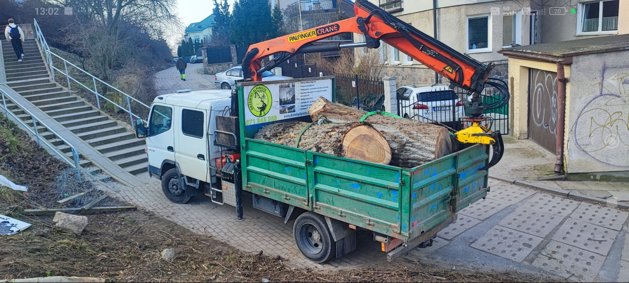 Ciężarówka z dźwigiem załadowana pociętymi pniami drzew, zaparkowana obok schodów z rampą. Widoczny budynek w tle i chodnik z płyt. Usługa wycinki drzew.