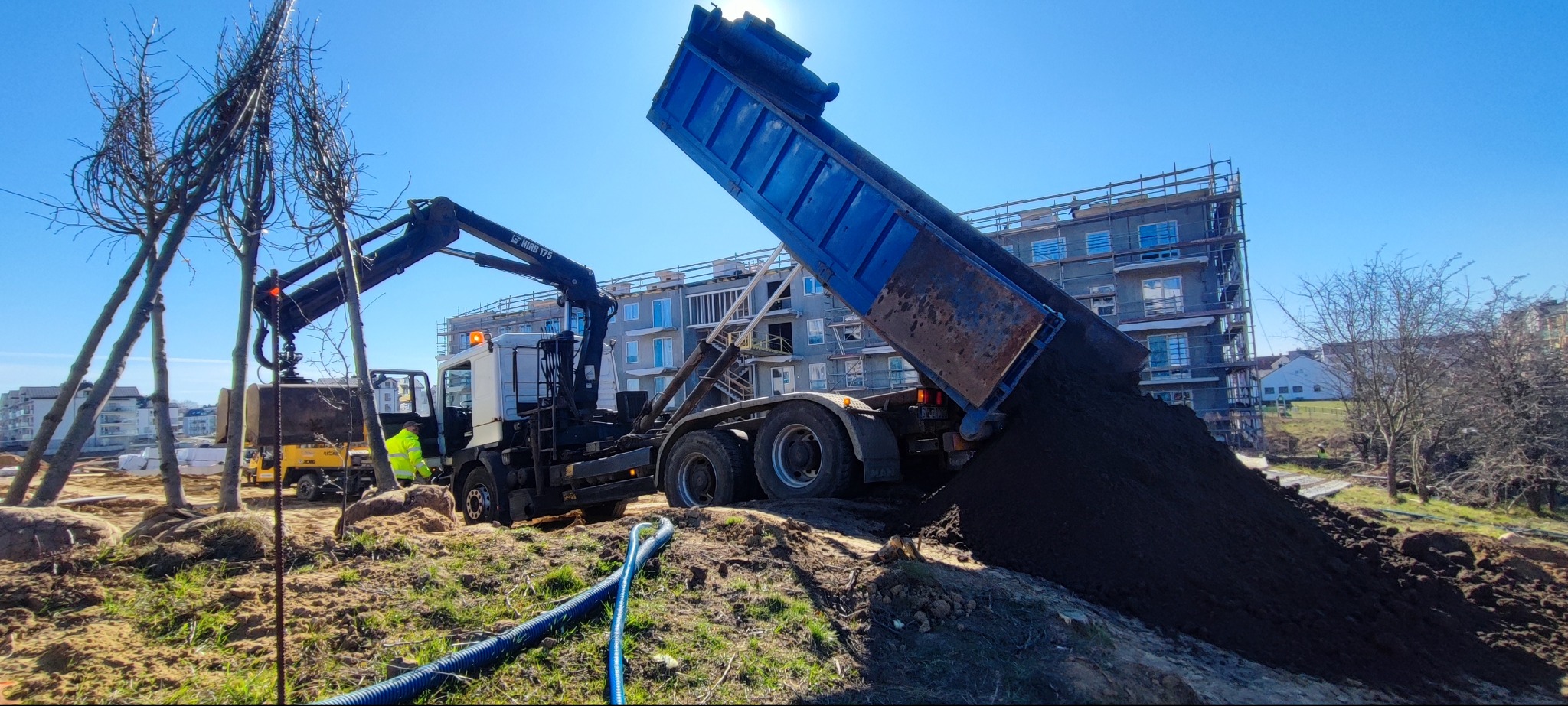 Ciężarówka z wywrotką wysypuje ziemię na plac budowy. W tle budynek w rusztowaniach i posadzone drzewa. Prace ziemne w słoneczny dzień. Widoczna niebieska rura.