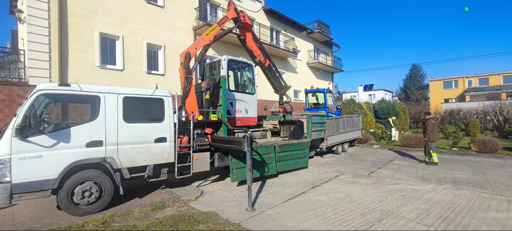 Ciężarówka z dźwigiem Palfinger i przyczepą z koparką na tle budynku. Dwóch mężczyzn w ogrodzie. Wykorzystanie ciężkiego sprzętu w pracach ogrodowych.