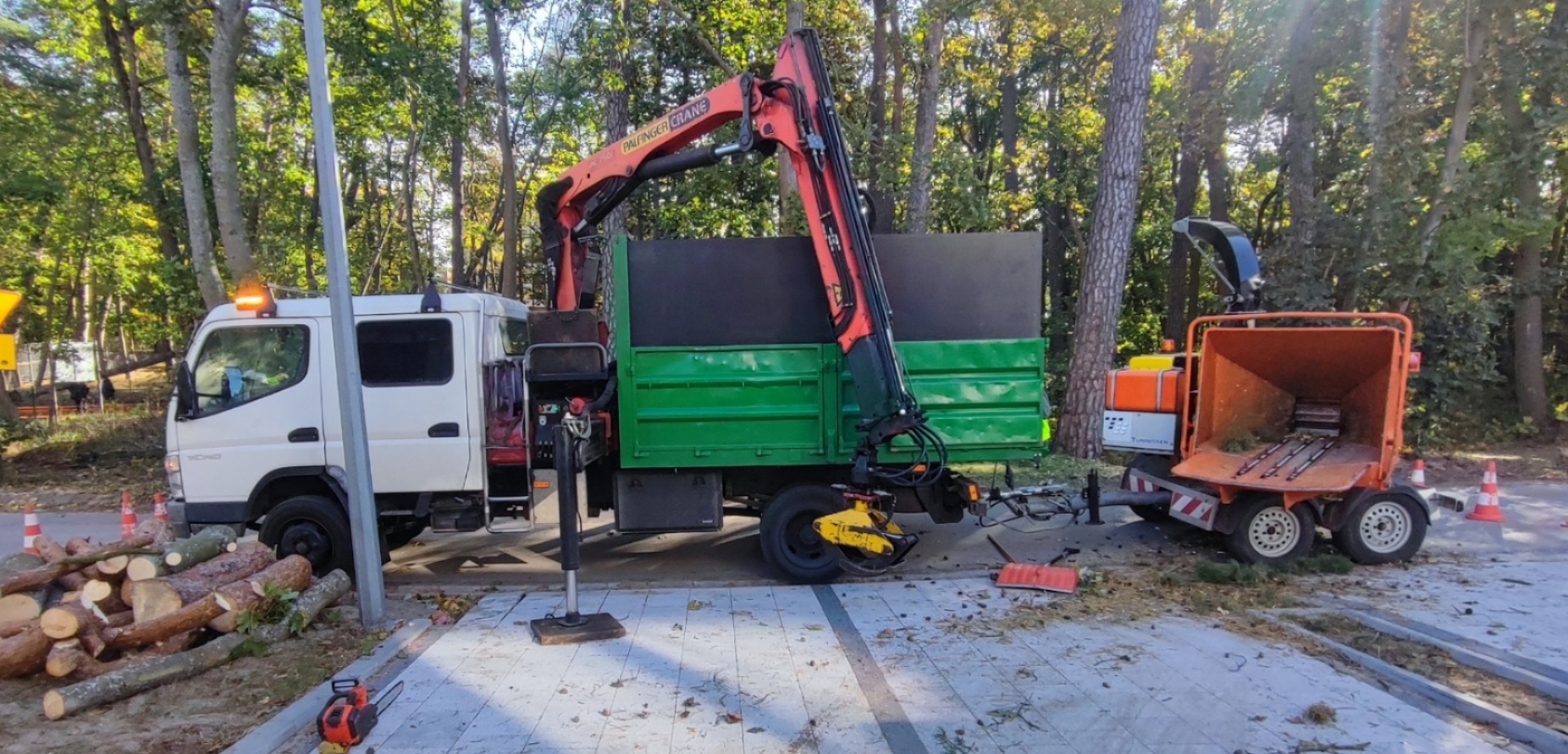 Ciężarówka z dźwigiem i rębakiem do gałęzi przy drodze, obok leżą pnie drzew. Sprzęt używany do wycinki drzew i utylizacji gałęzi. Praca w toku, widoczne ścięte pnie.