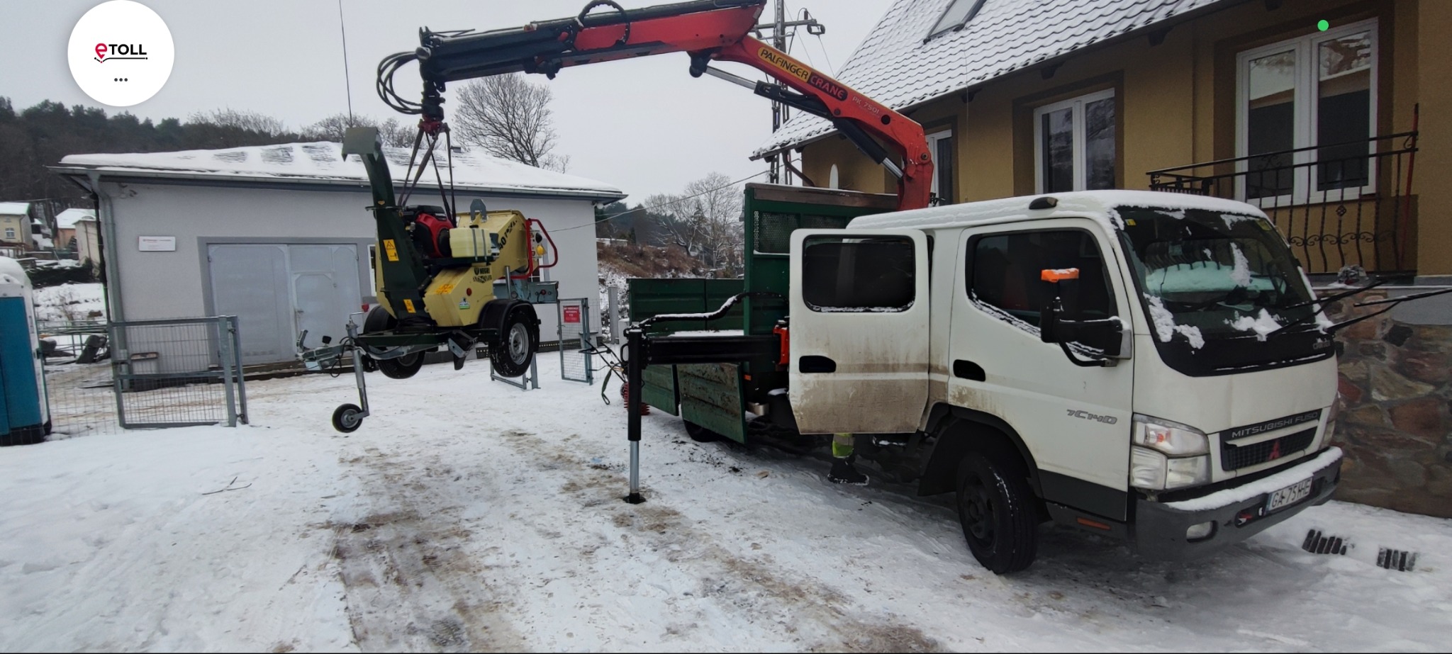 Ciężarówka z dźwigiem podnosi rozdrabniacz do gałęzi na tle zaśnieżonego terenu i zabudowań. Widoczny fragment elewacji budynku.