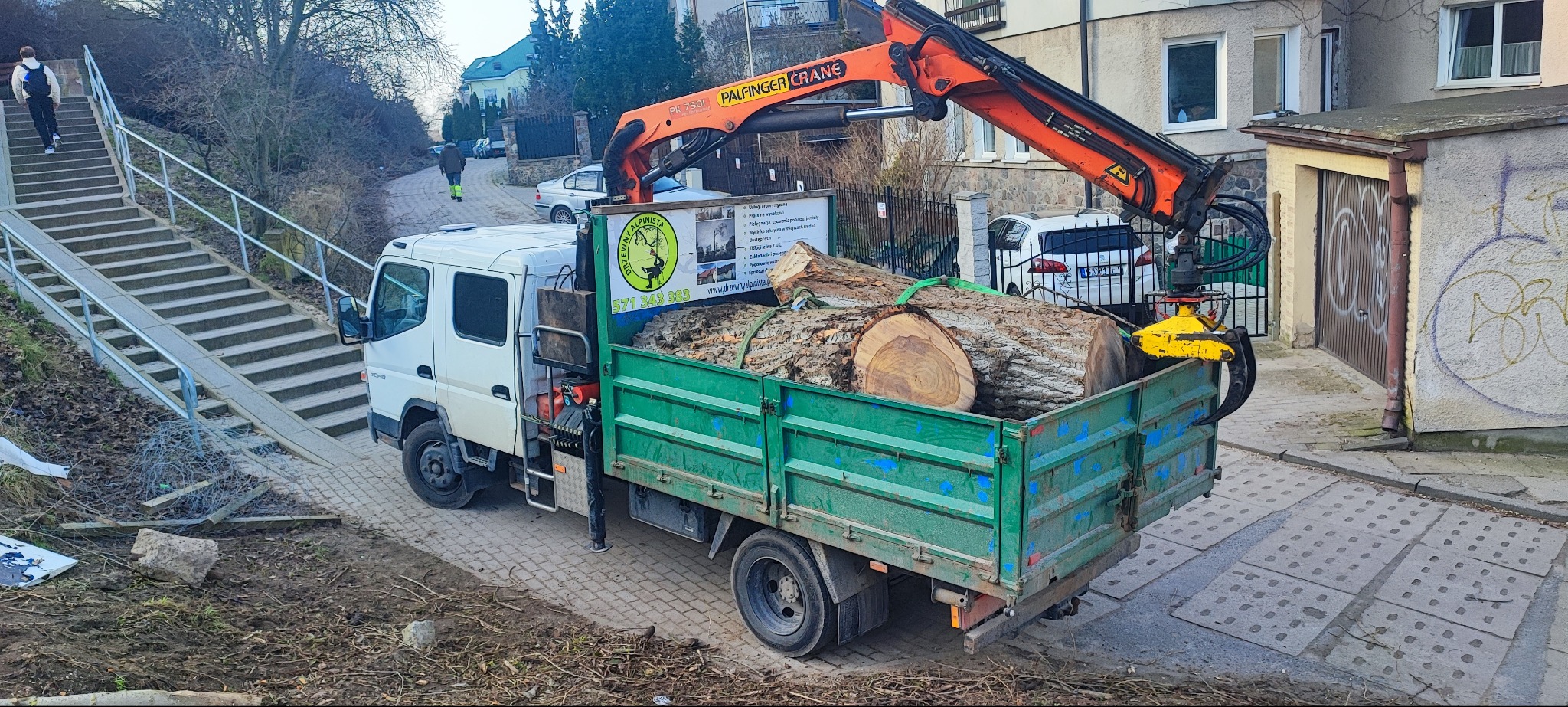 Ciężarówka z dźwigiem PALFINGER załadowana pociętymi pniami drzew, na tle budynków i schodów. Widoczny przekrój pni.