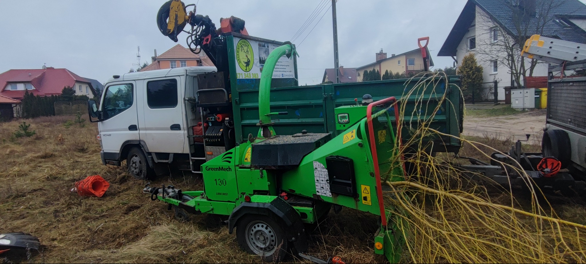 Mobilny rębak GreenMech 130 Arborist przy białej ciężarówce z dźwigiem, przygotowany do pracy na tle domów. Widoczne gałęzie i pomarańczowy pachołek.