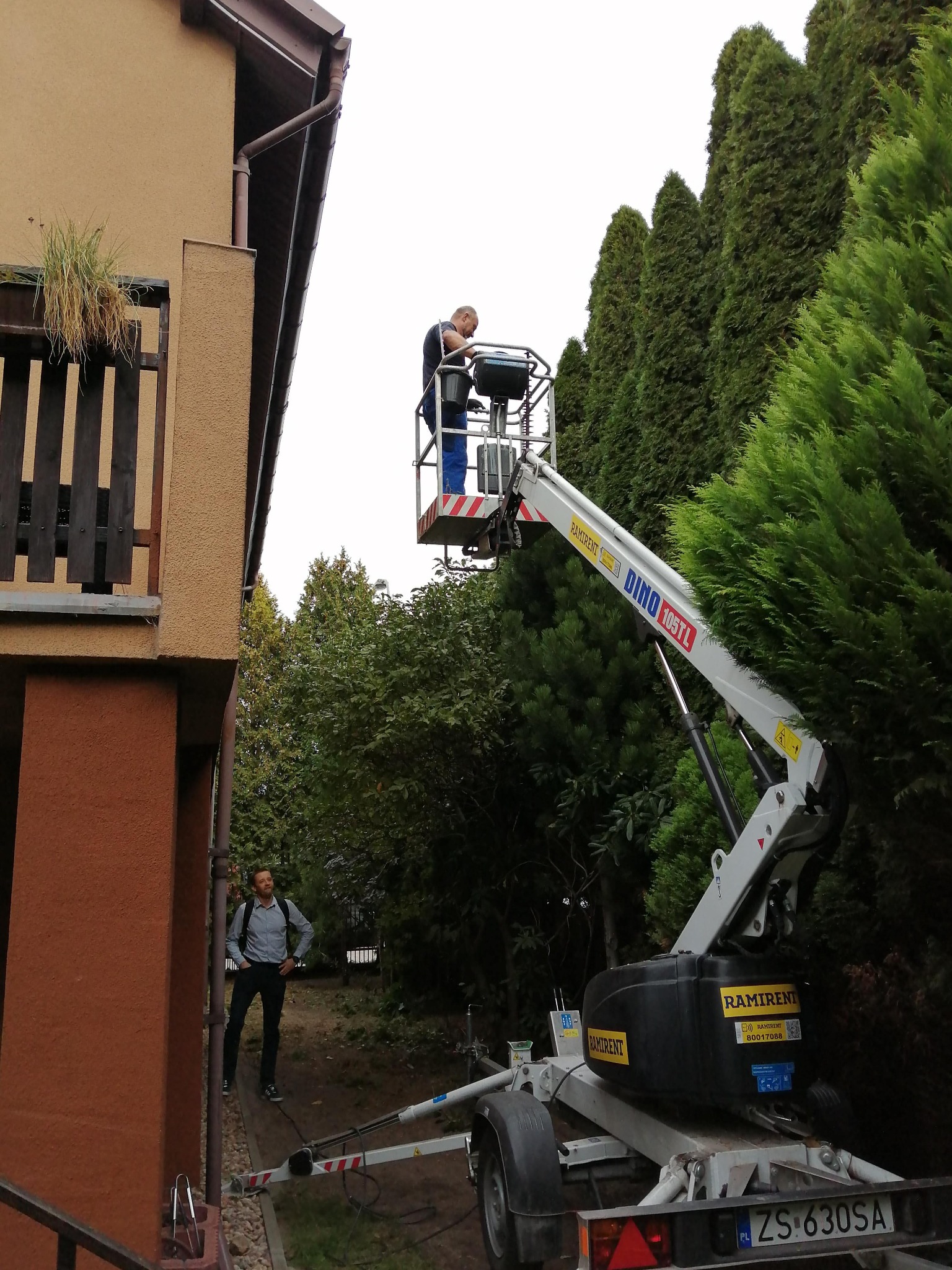 Pracownik na podnośniku koszowym DINO 105TL przy budynku, prawdopodobnie wykonuje prace pielęgnacyjne drzew, drugi mężczyzna obserwuje z dołu.