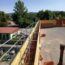 Zbrojenie stropu w drewnianym szalunku z widocznymi prętami i cegłami, na tle zieleni i błękitnego nieba. Widok z góry na konstrukcję w trakcie budowy.