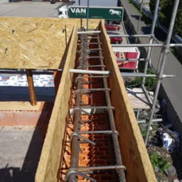 Zbrojenie w szalunku z desek na murze z czerwonej cegły, widok z góry. W tle budynki i zieleń. Elementy rusztowania widoczne po prawej stronie.