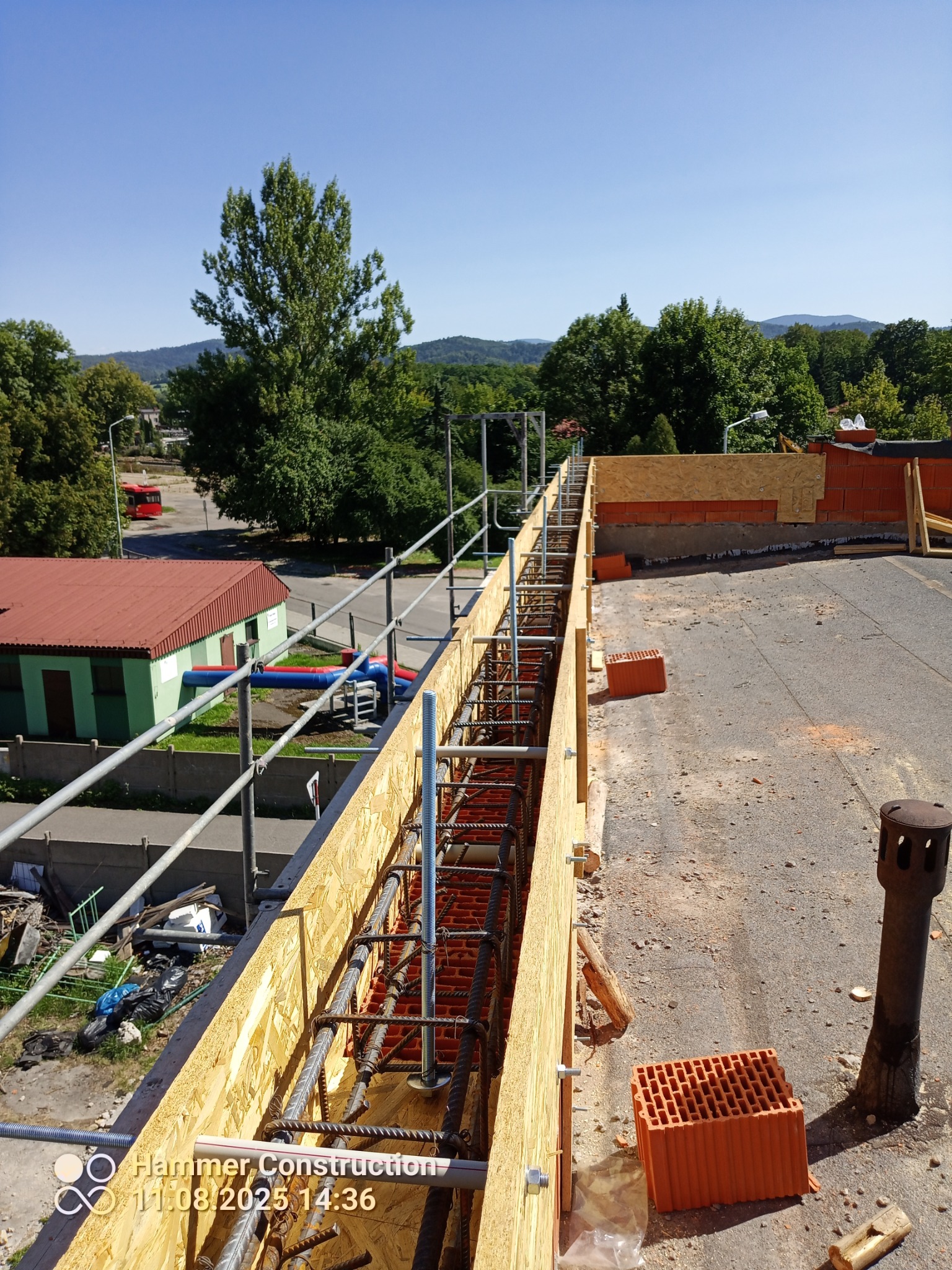 Zbrojenie stropu w drewnianym szalunku z widocznymi prętami i cegłami, na tle zieleni i błękitnego nieba. Widok z góry na konstrukcję w trakcie budowy.