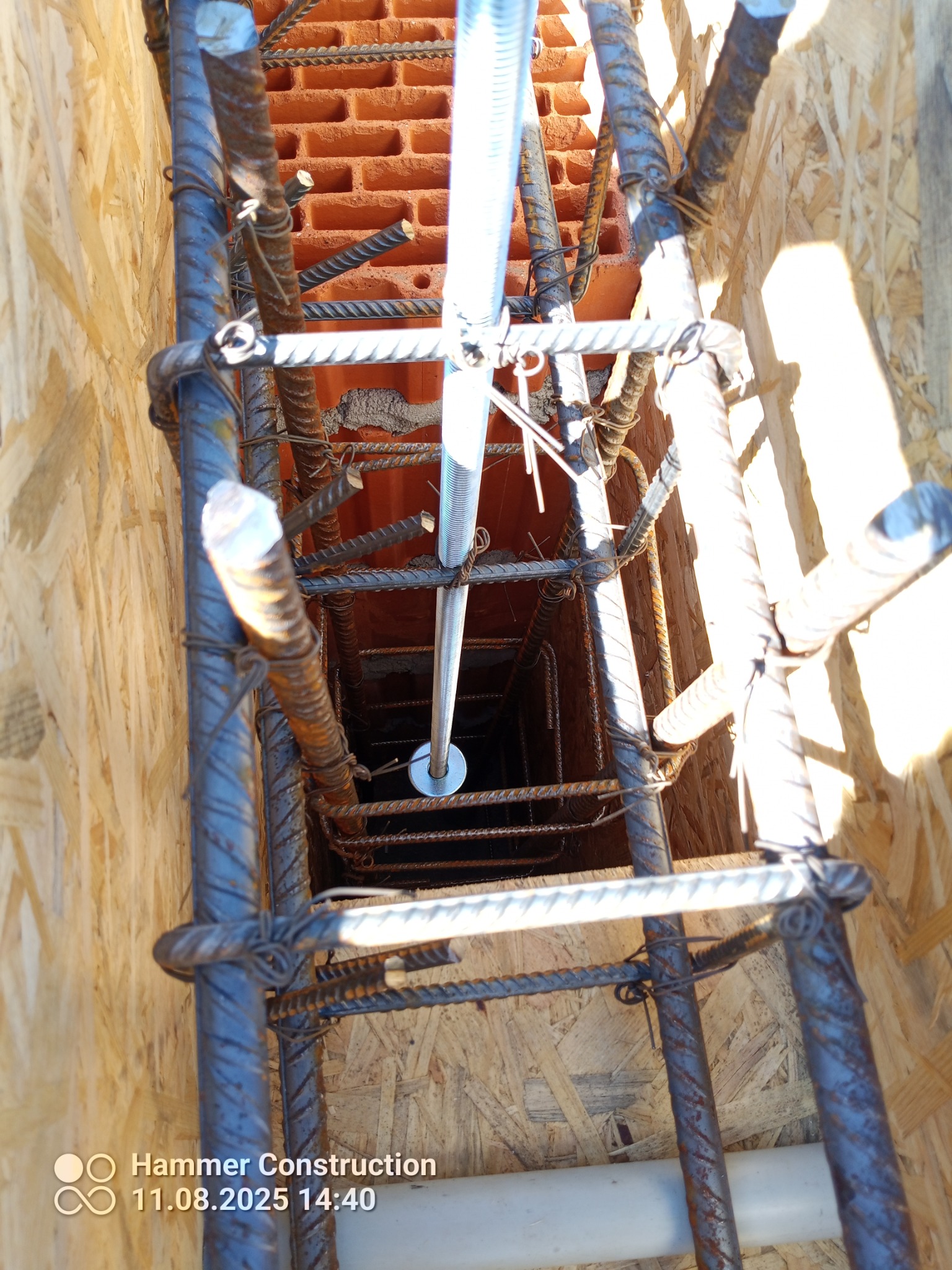 Zbrojenie fundamentu z widocznymi prętami stalowymi, deskowaniem z płyty OSB i fragmentem ściany z czerwonej cegły. Widok z góry na konstrukcję.