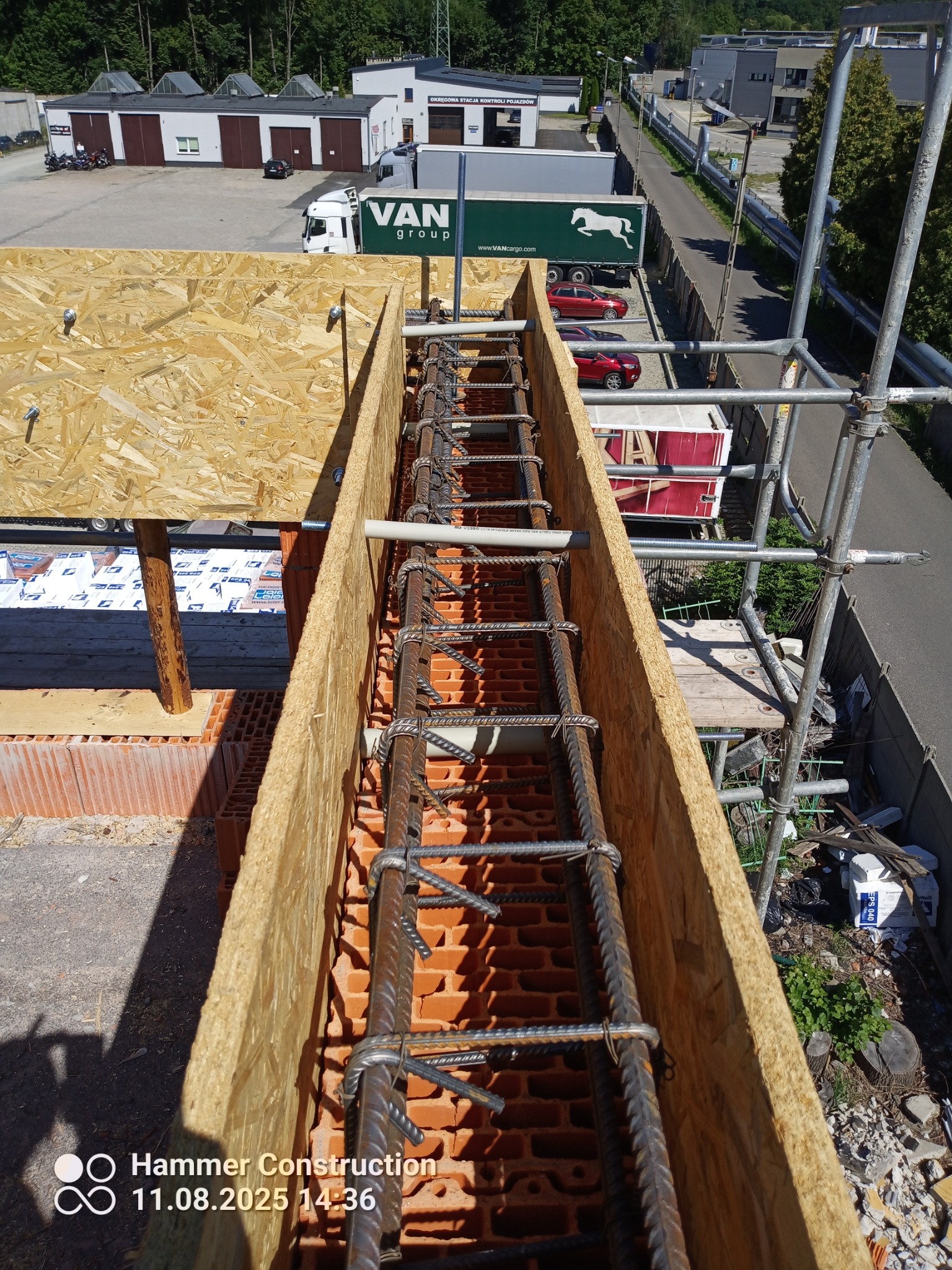 Zbrojenie w szalunku z desek na murze z czerwonej cegły, widok z góry. W tle budynki i zieleń. Elementy rusztowania widoczne po prawej stronie.