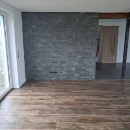 Salon po remoncie: drewnopodobne panele na podłodze, szara ściana z kamienia dekoracyjnego z gniazdkami, okno z białymi ramami. Widok na sąsiednie pomieszczenie.