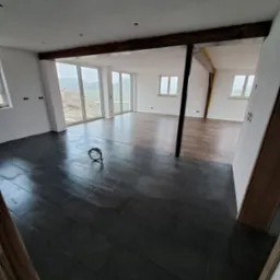 Nowoczesne wnętrze z ciemną, błyszczącą podłogą w jodełkę, drewnianymi belkami stropowymi i dużymi oknami z widokiem na zewnątrz. Częściowo ułożona podłoga w sąsiednim pomieszczeniu.