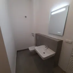 Nowoczesna łazienka z podświetlanym lustrem, umywalką nablatową i szarą zabudową. Minimalistyczny design, szare płytki podłogowe. Widoczny fragment grzejnika.