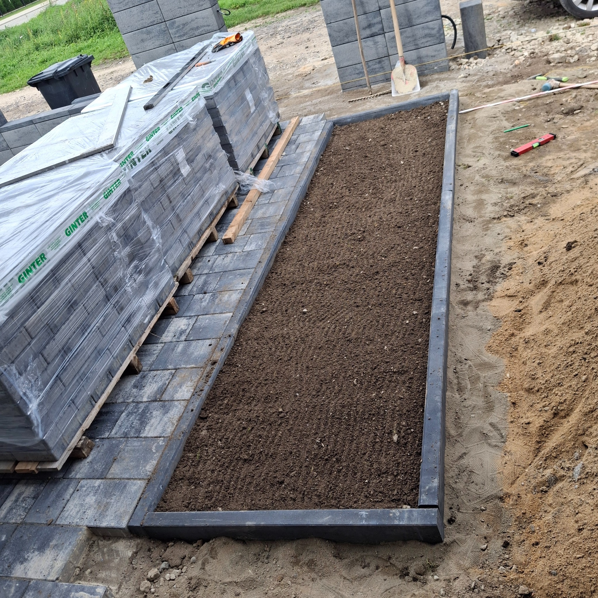 Przygotowanie rabaty kwiatowej z obrzeżami z kostki brukowej, obok paleta kostki w folii. Widoczne narzędzia i poziomica.