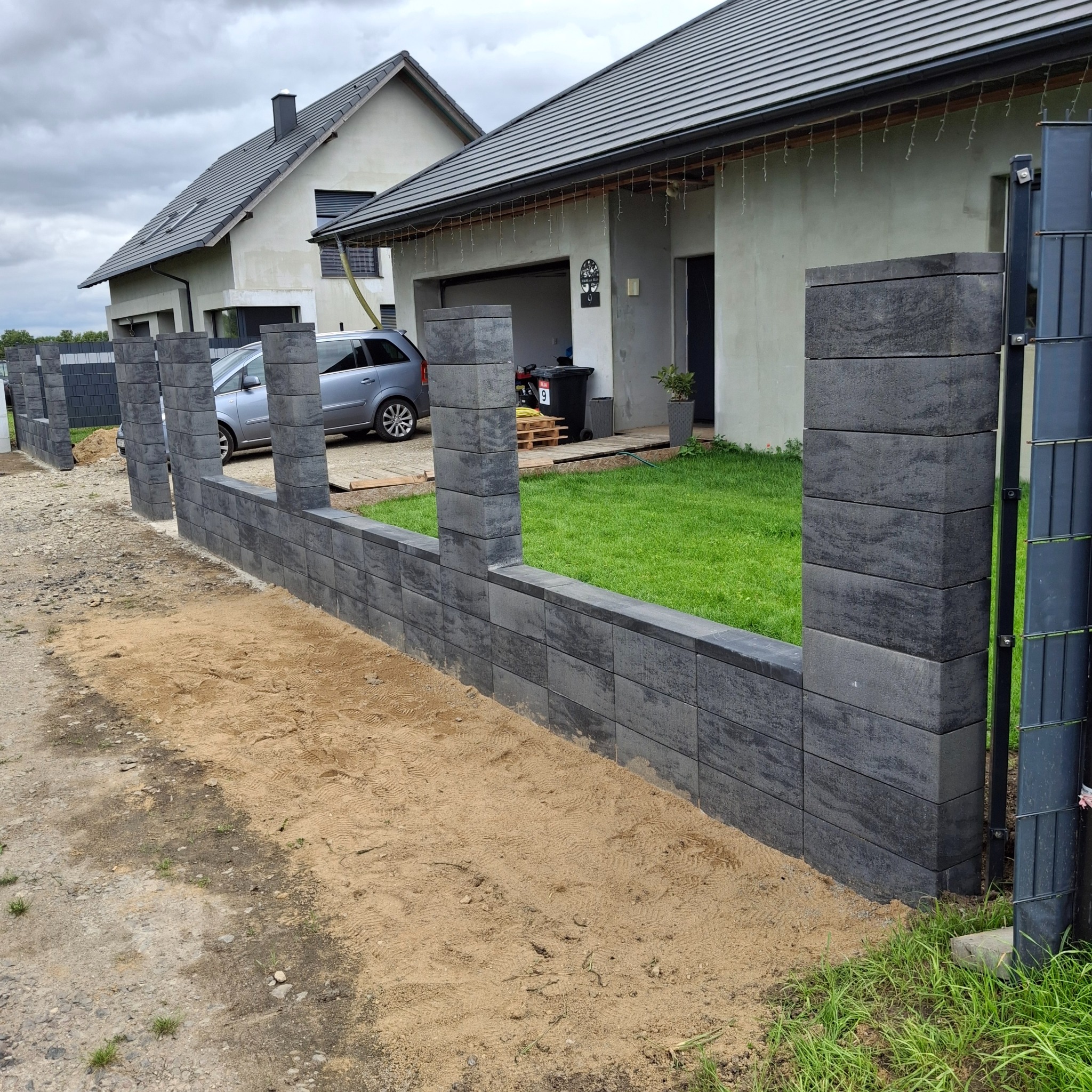 Nowoczesny, częściowo ukończony mur z szarej kostki, z widocznymi słupkami, na tle domu z szarą dachówką i zielonego trawnika. Widoczny piasek sugeruje trwające prace.