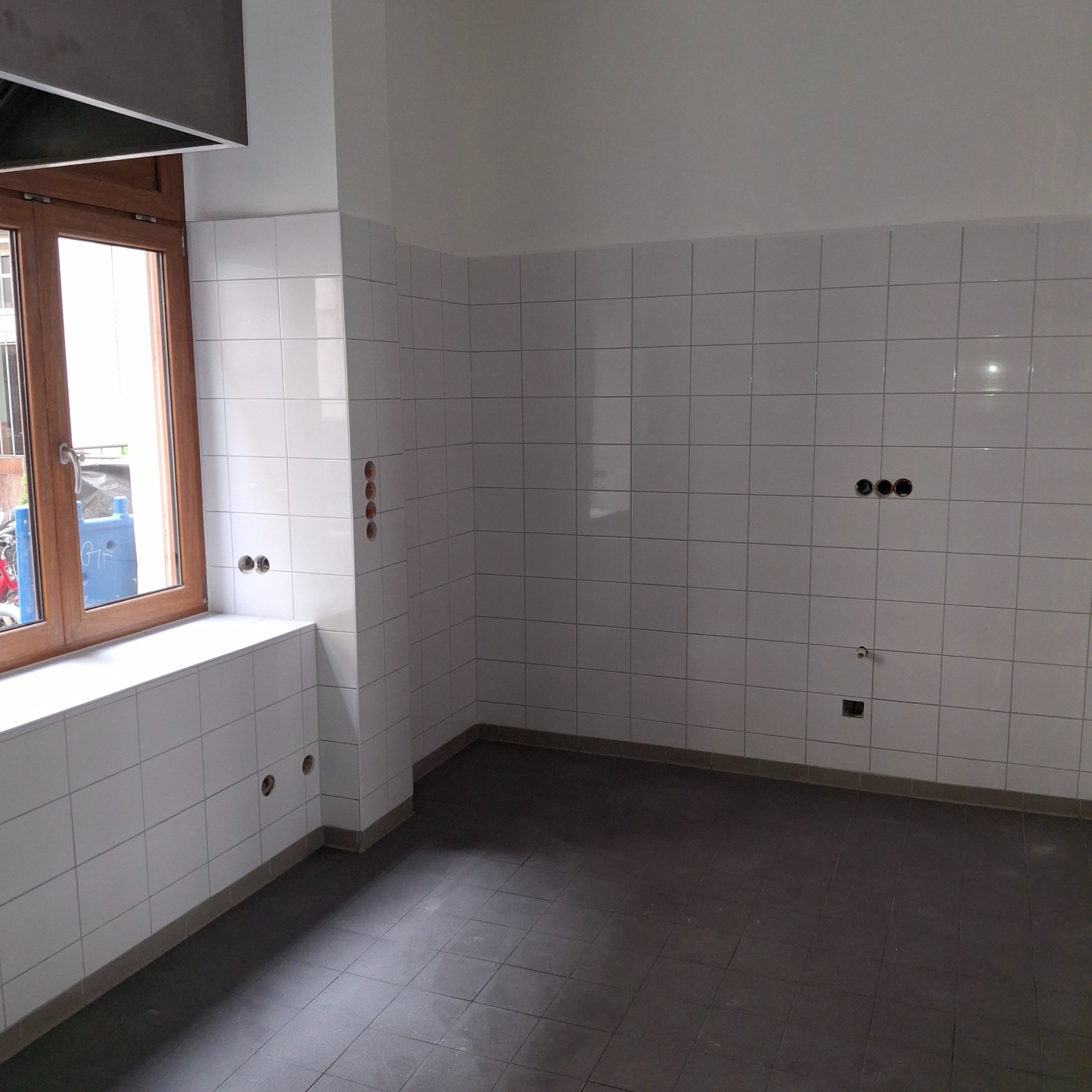 Wnętrze po wykończeniu: białe płytki na ścianach, ciemna podłoga z płytek. Widoczne gniazdka elektryczne i fragment okna z drewnianą ramą. Minimalistyczny design.