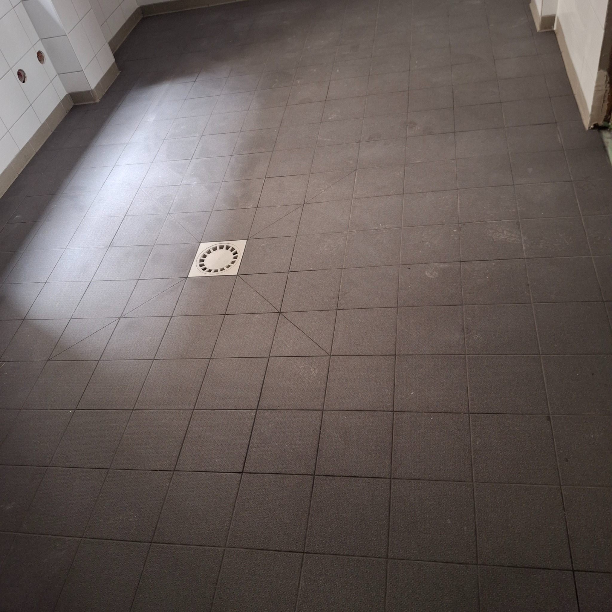 Ciemne, kwadratowe płytki podłogowe z odpływem w centrum, ułożone w łazience. Widoczne białe płytki na ścianach i fragment niedokończonego narożnika.