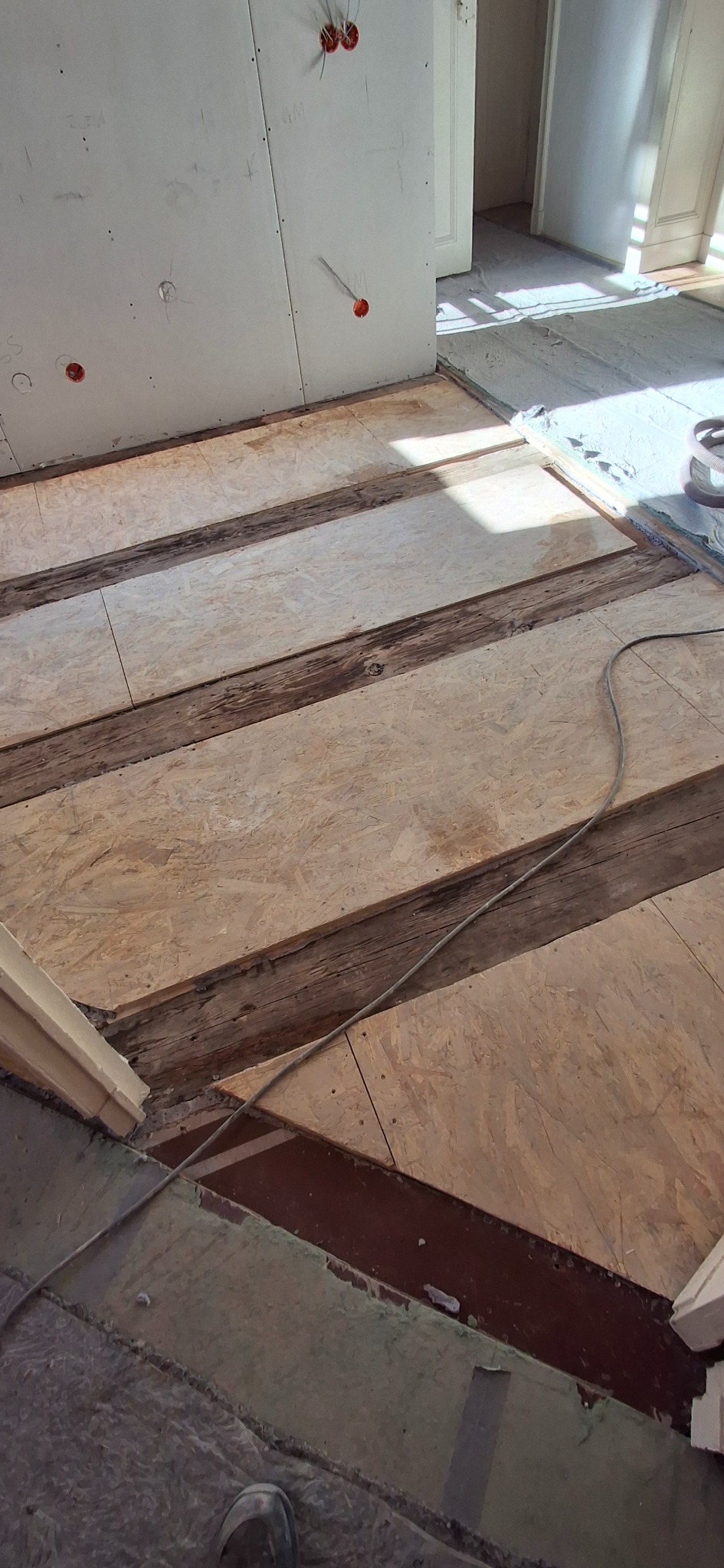 Przygotowanie podłoża pod panele: deski drewniane z płytami OSB, widoczne kable elektryczne w ścianie. Etap prac remontowych w jasnym pomieszczeniu.