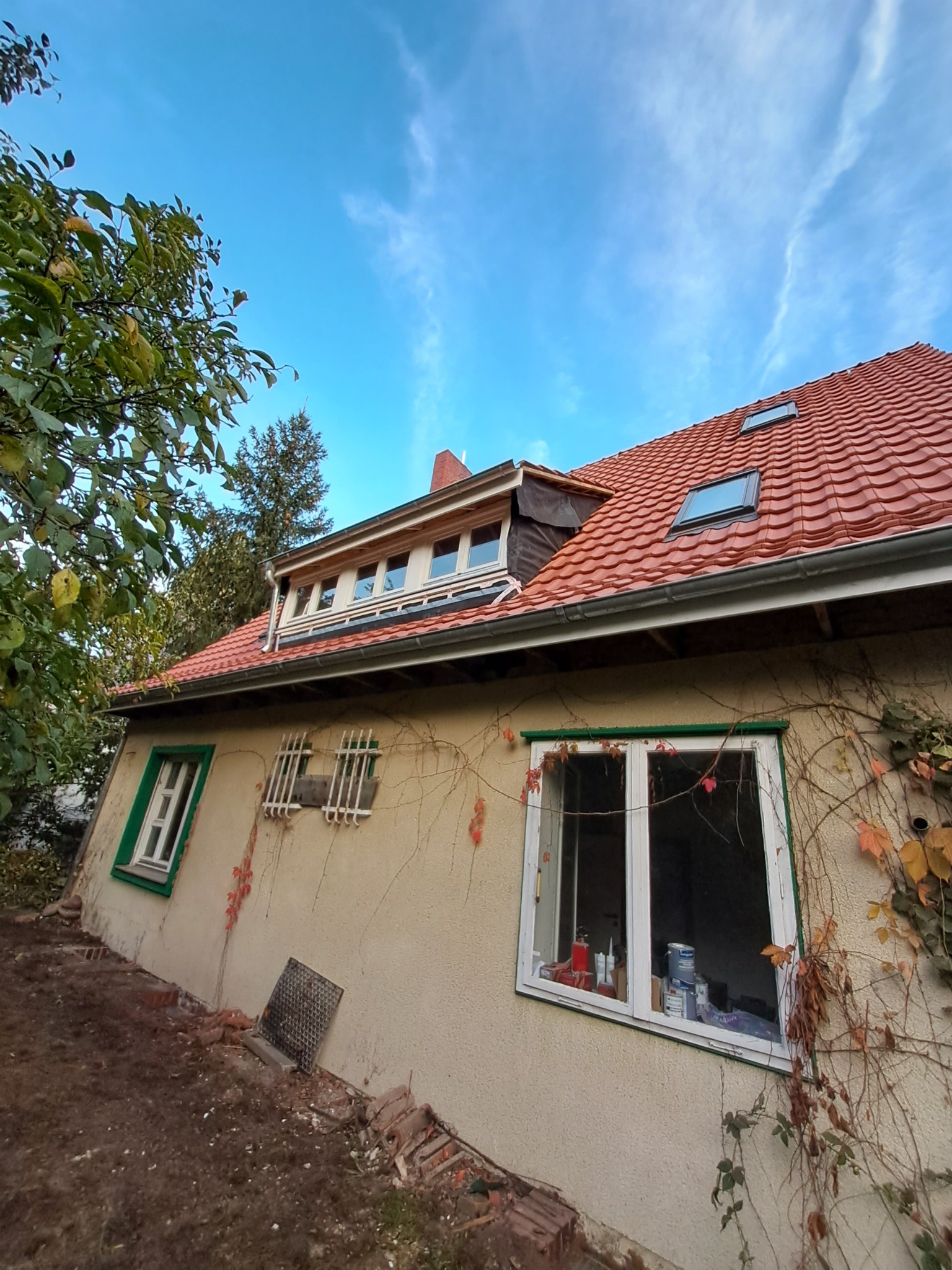 Elewacja budynku z czerwonym dachem i oknami z zielonymi ramami, obrośnięta bluszczem. Widoczne okna dachowe i fragment niezagospodarowanego terenu.