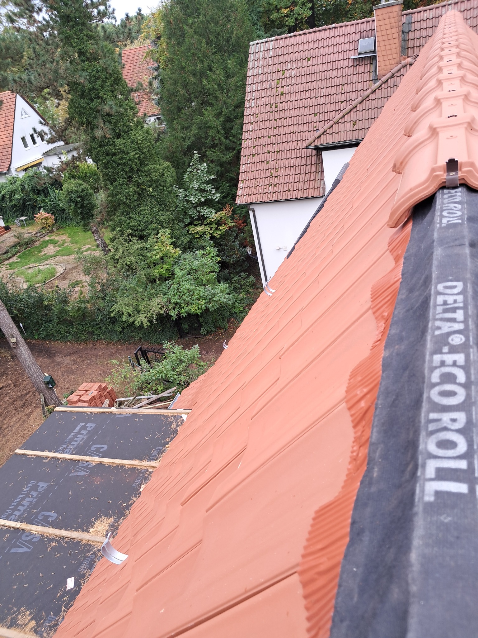 Czerwona dachówka na nowym dachu w trakcie budowy, widok z góry pod kątem. Widoczne materiały izolacyjne i fragmenty domów w tle.