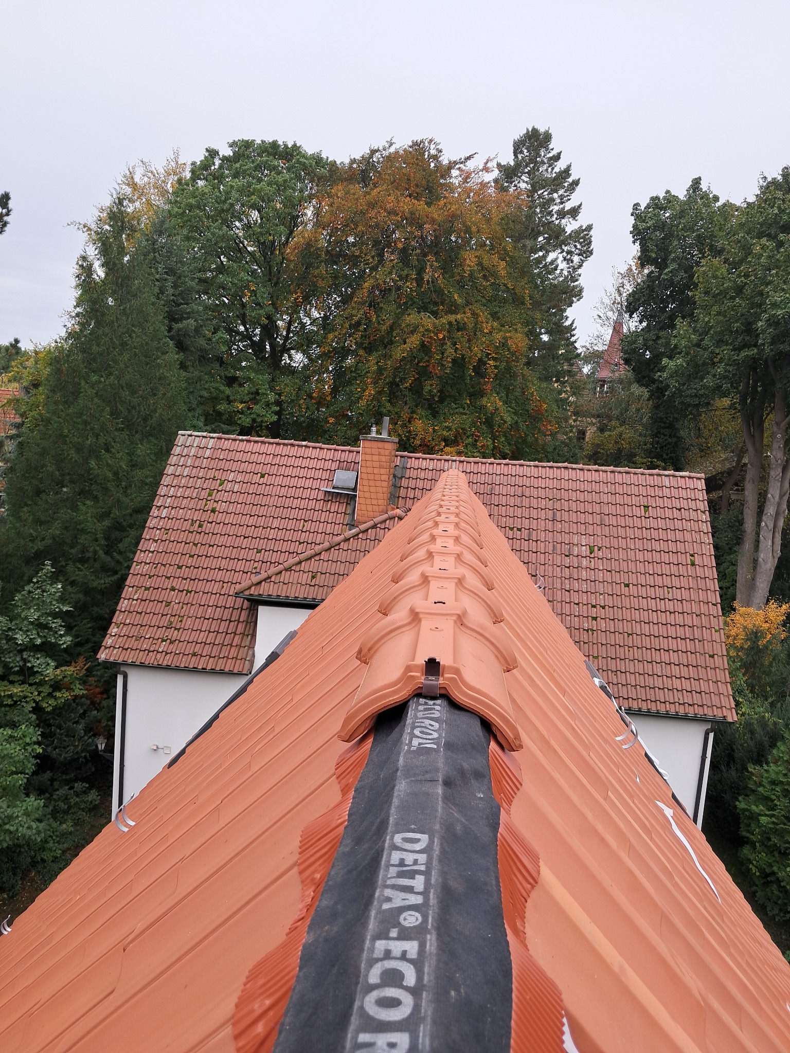 Czerwona dachówka ceramiczna z widoczną membraną Delta Eco Pol na dachu domu, z zielenią drzew w tle i fragmentem sąsiedniego budynku.