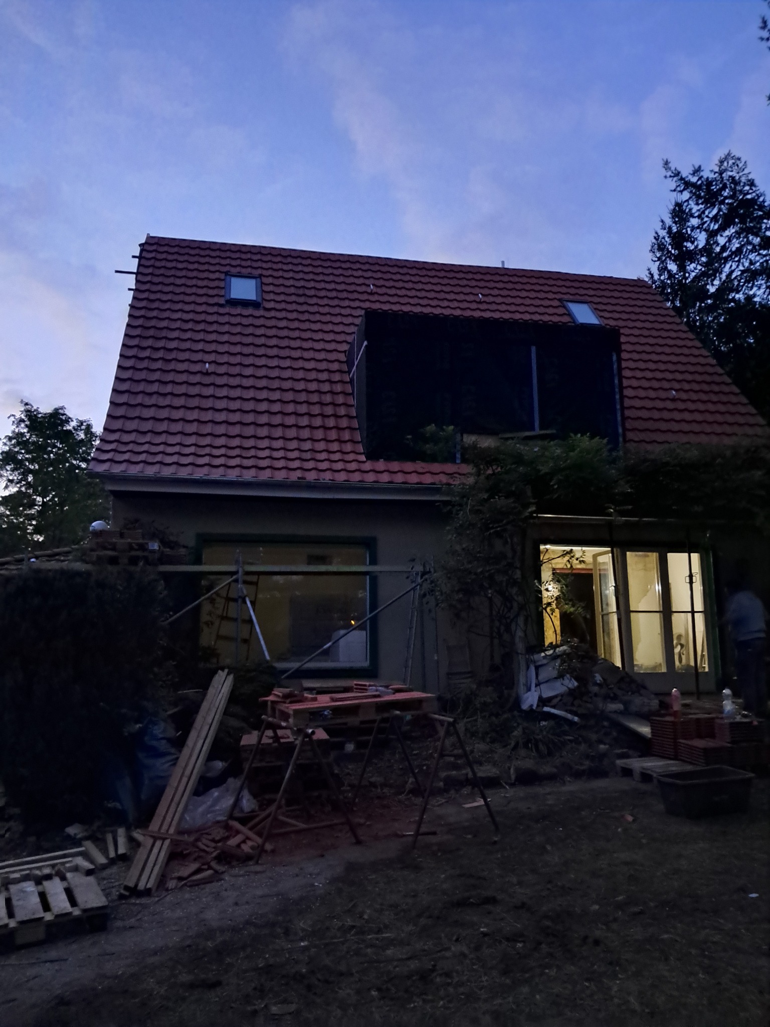 Dom jednorodzinny w trakcie remontu elewacji i dachu, widoczne rusztowania, palety z dachówkami i materiały budowlane. Częściowo odsłonięta konstrukcja dachu.