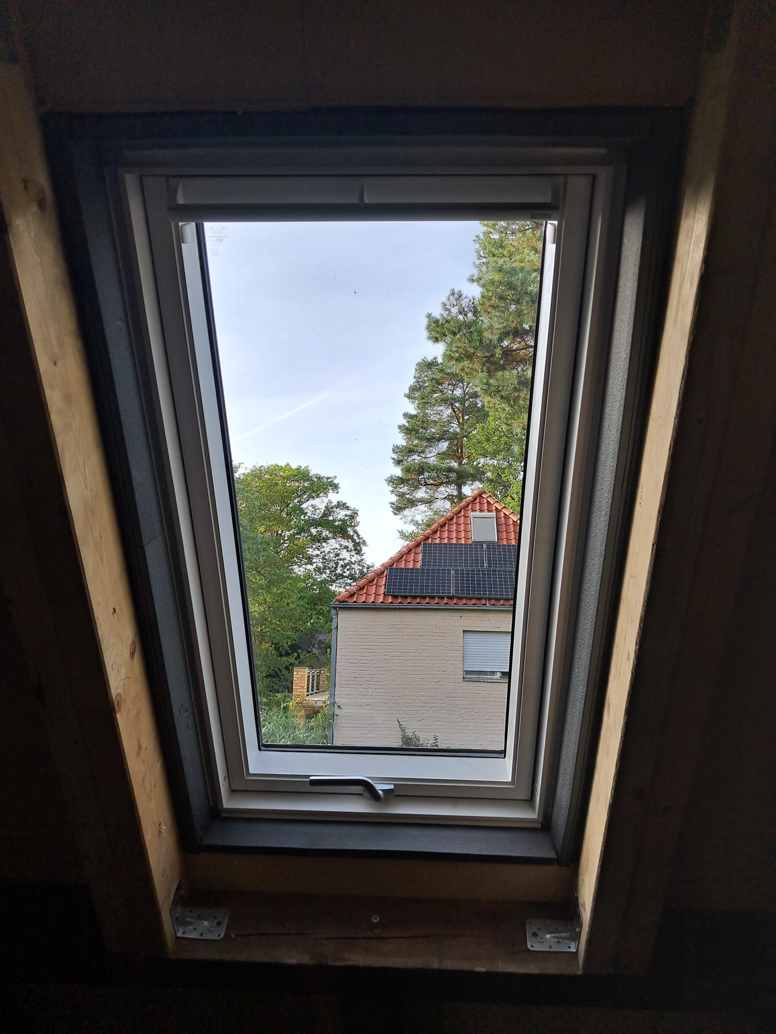 Okno dachowe z widokiem na dom z panelami słonecznymi i drzewa. Drewniana rama okna w trakcie montażu, widoczne elementy konstrukcyjne.