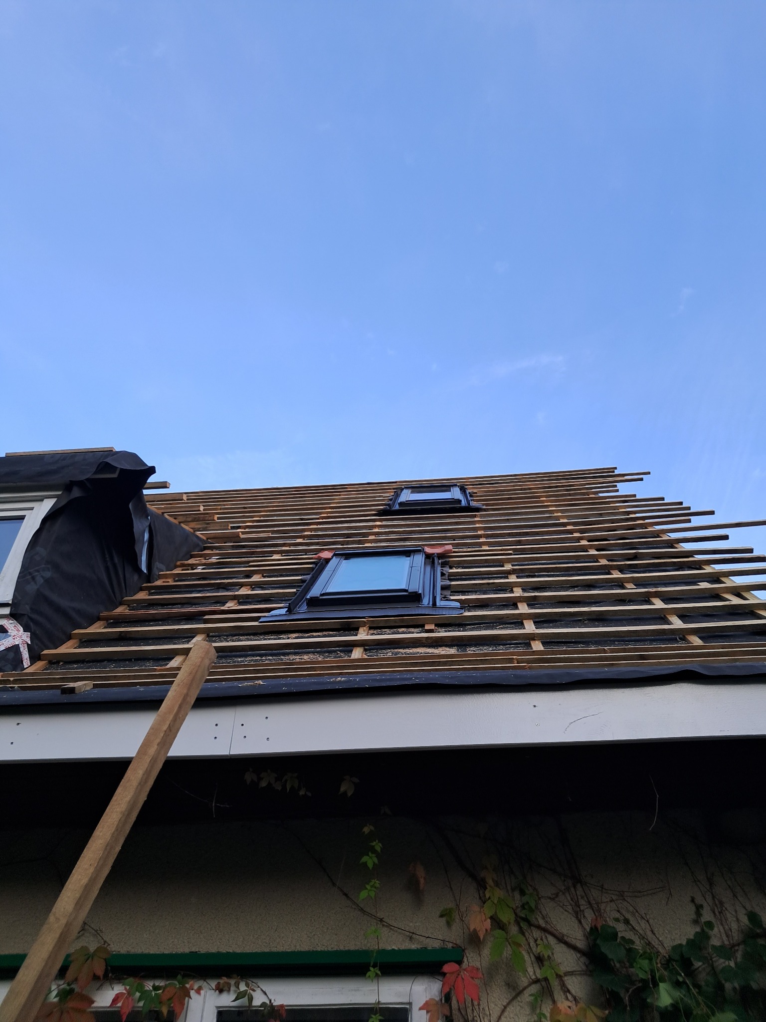 Montaż łat na dachu z oknami połaciowymi, widok z dołu, częściowo pokryty membraną. Elementy drewniane i okna na tle jasnego nieba.